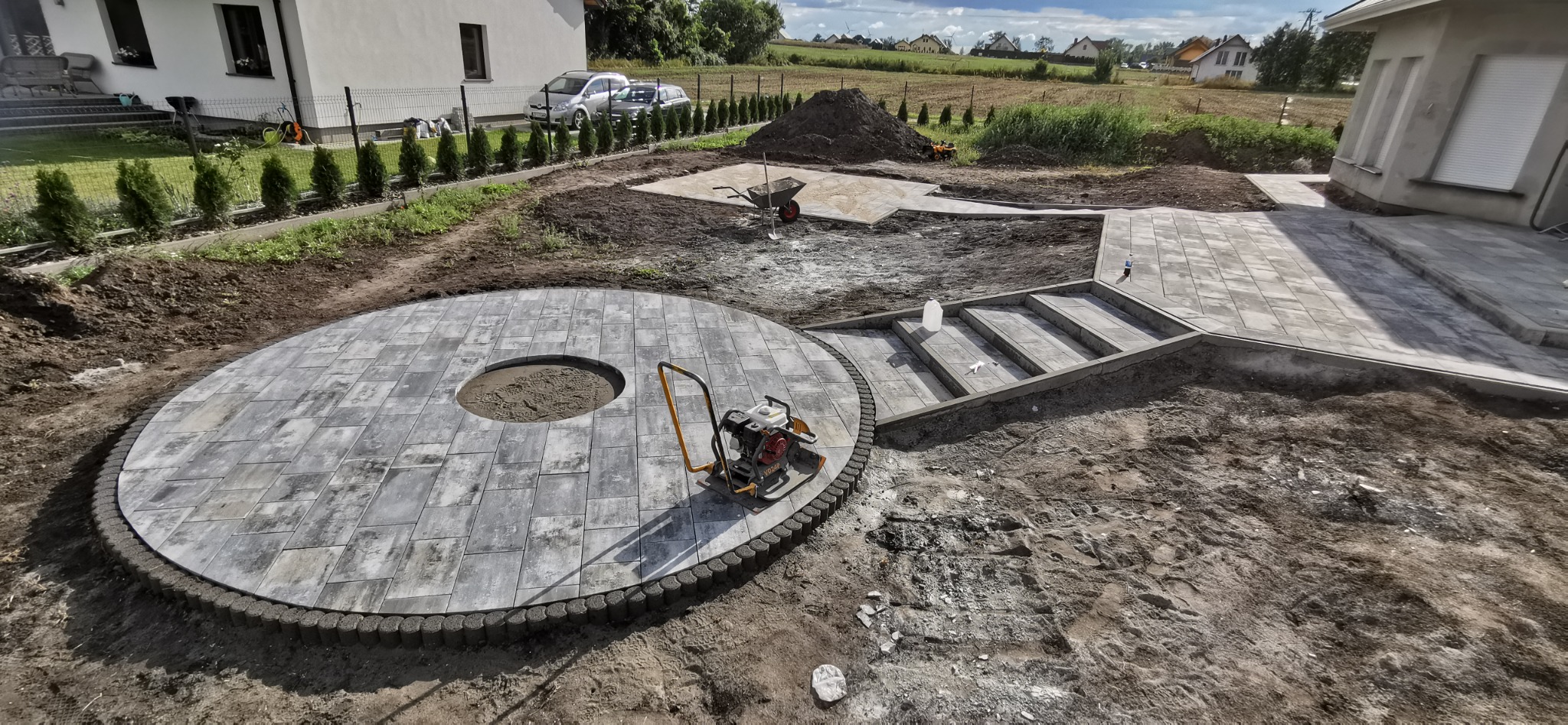 Układanie szarej kostki brukowej w okrąg wokół przyszłego paleniska, widoczna zagęszczarka spalinowa, schody z kostki brukowej oraz fragment ścieżki w trakcie realizacji.