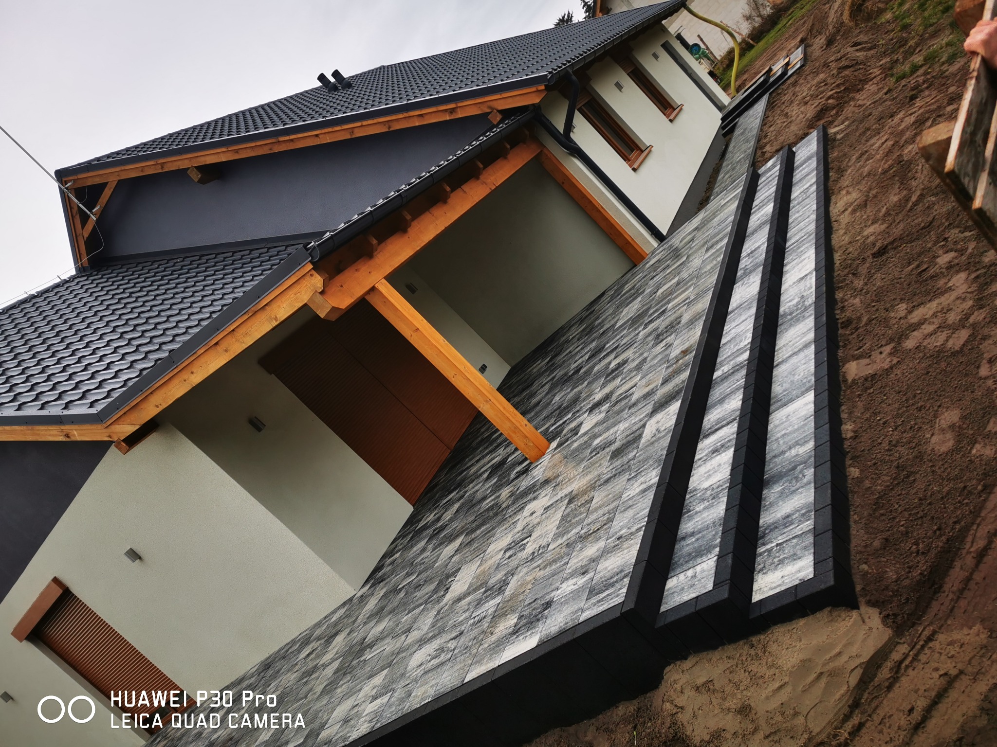 Układanie szarej kostki na podjeździe wzdłuż domu z ciemną dachówką, widoczne czarne obrzeża kostki i fragmenty ziemi.