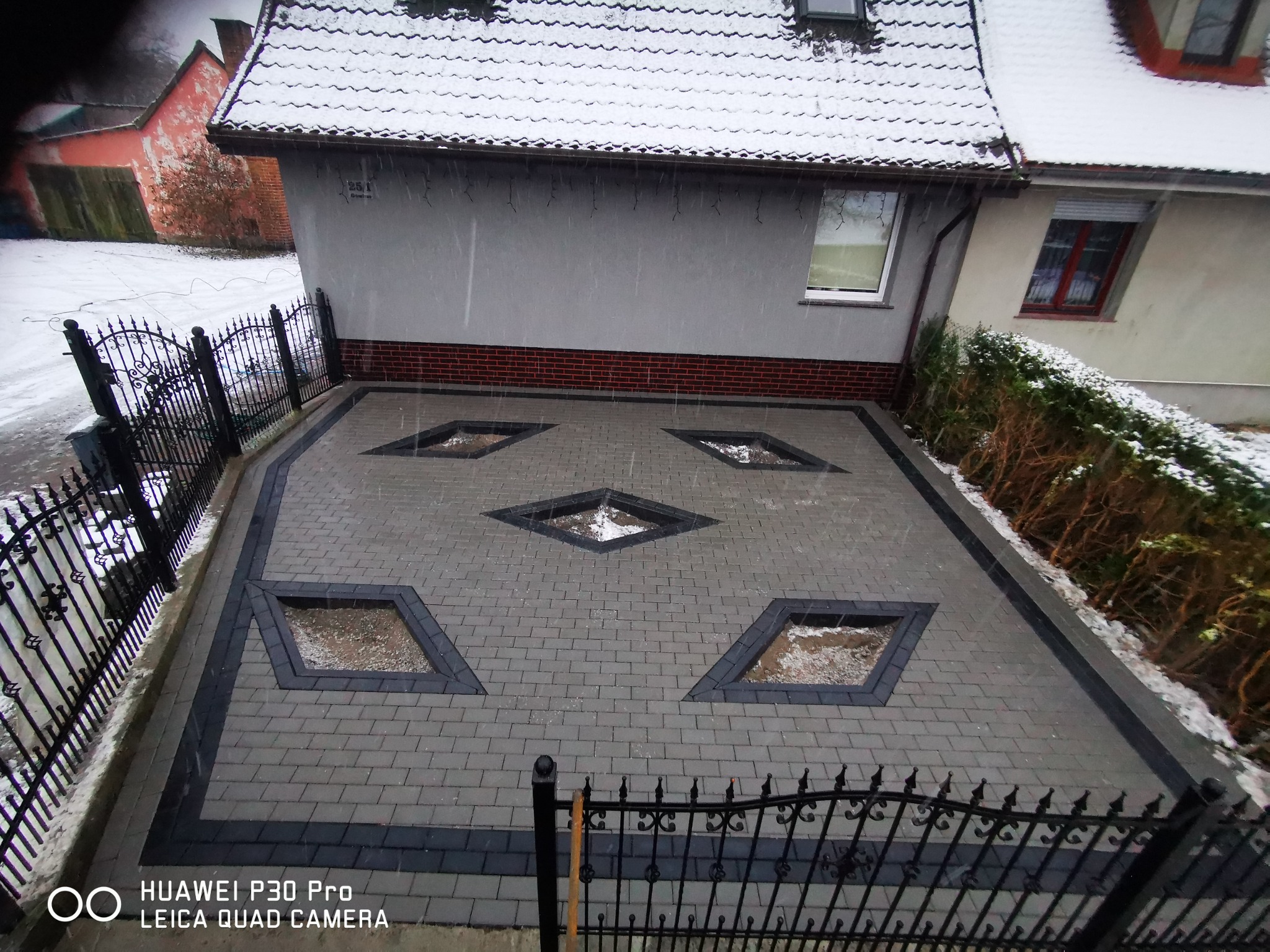 Ułożona szara kostka brukowa z czarnymi obramowaniami i romboidalnymi wgłębieniami wypełnionymi wodą, widok z góry przez ozdobne metalowe ogrodzenie w zimowy, pochmurny dzień.