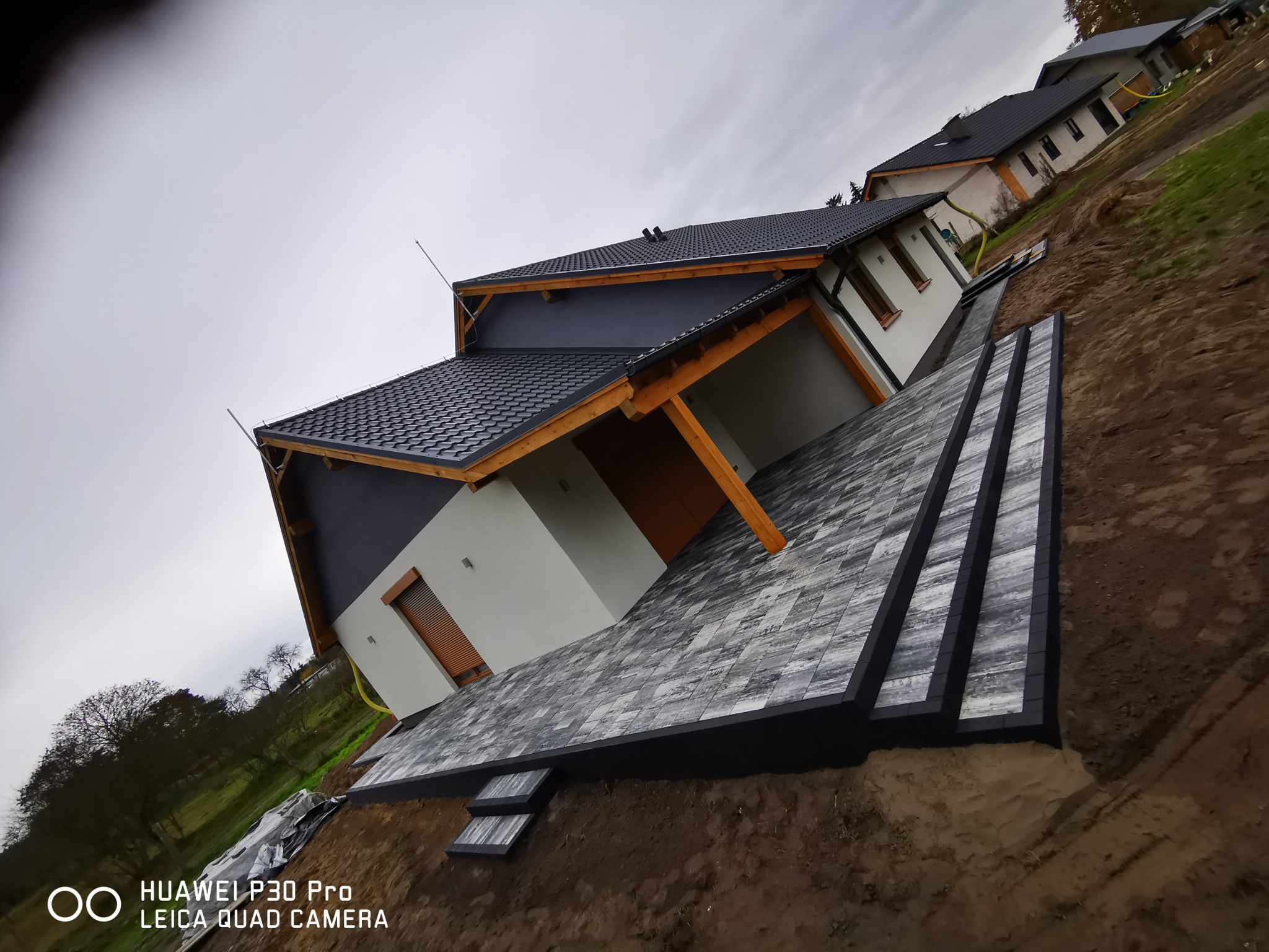 Nowoczesny dom z szaro-białą elewacją i tarasem z kostki brukowej o geometrycznym wzorze, otoczony ziemią i innymi domami w tle.