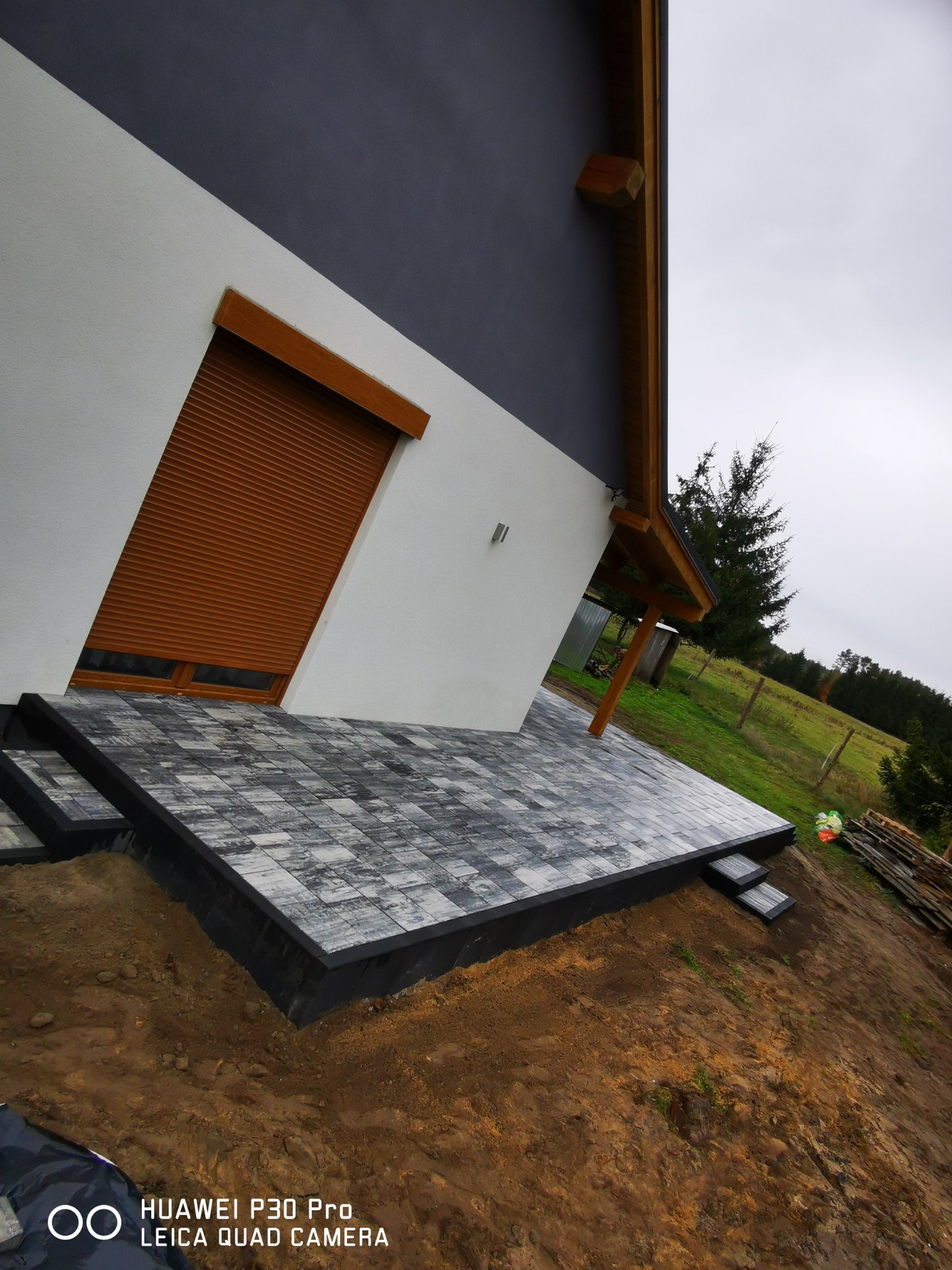 Nowoczesny taras z szarej kostki brukowej ułożony przy domu z brązowymi roletami okiennymi, widok z perspektywy ziemi, w tle zieleń.