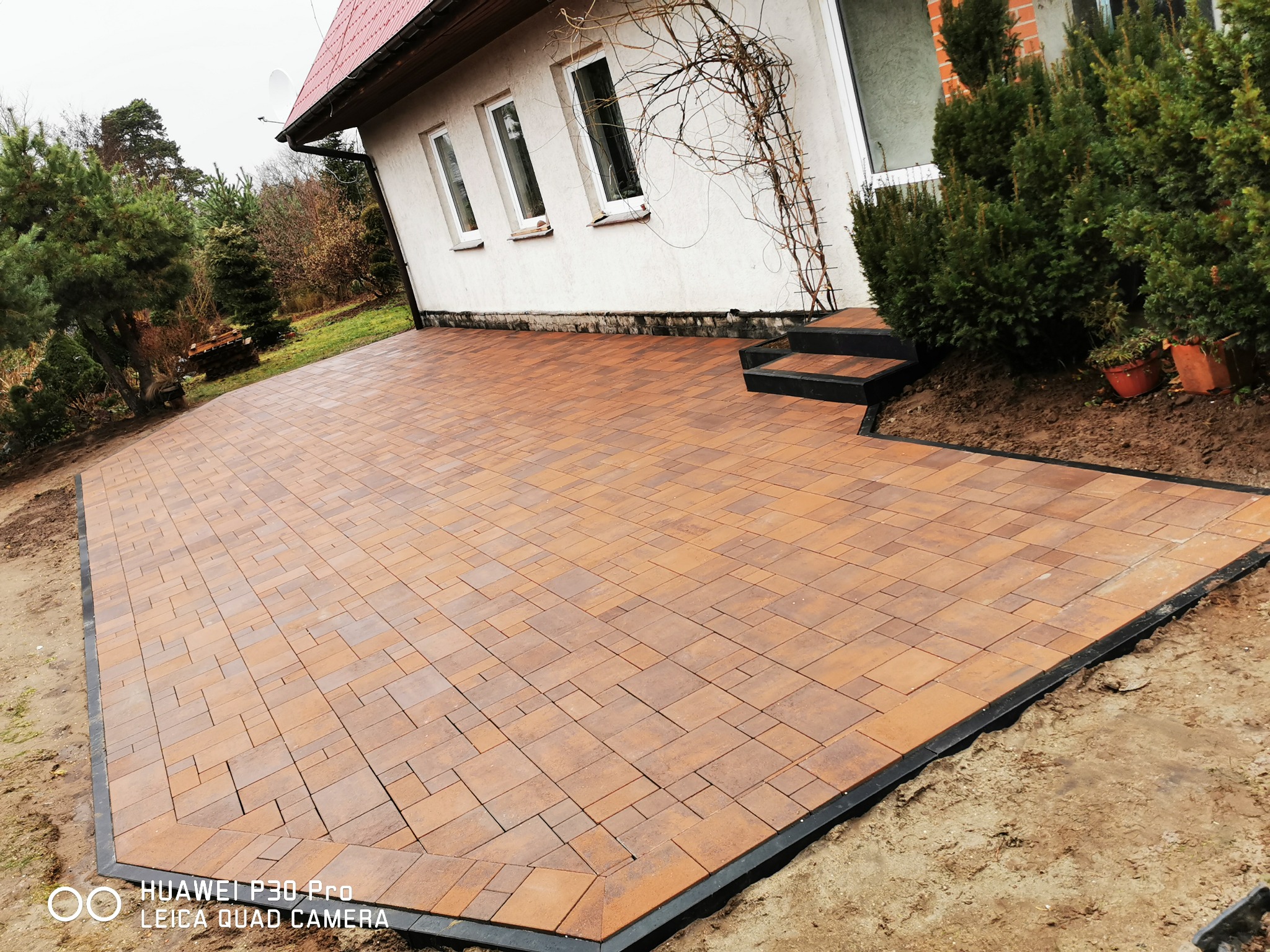 Nowo ułożony, geometryczny wzór z brązowej kostki brukowej otoczony czarnym obrzeżem, przylegający do białego domu z trzema oknami i schodami wejściowymi, widok z poziomu gruntu.