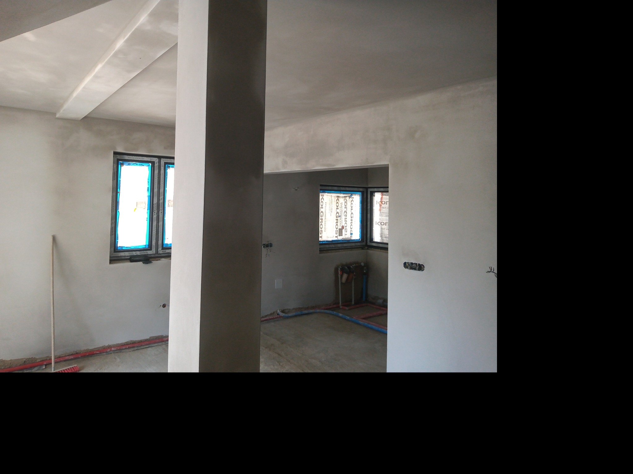 Surowe wnętrze domu w trakcie prac wykończeniowych: ściany i sufit pokryte świeżym, jasnoszarym tynkiem, widoczne rury instalacji grzewczej w podłodze, okna zabezpieczone folią ochronną, brak...