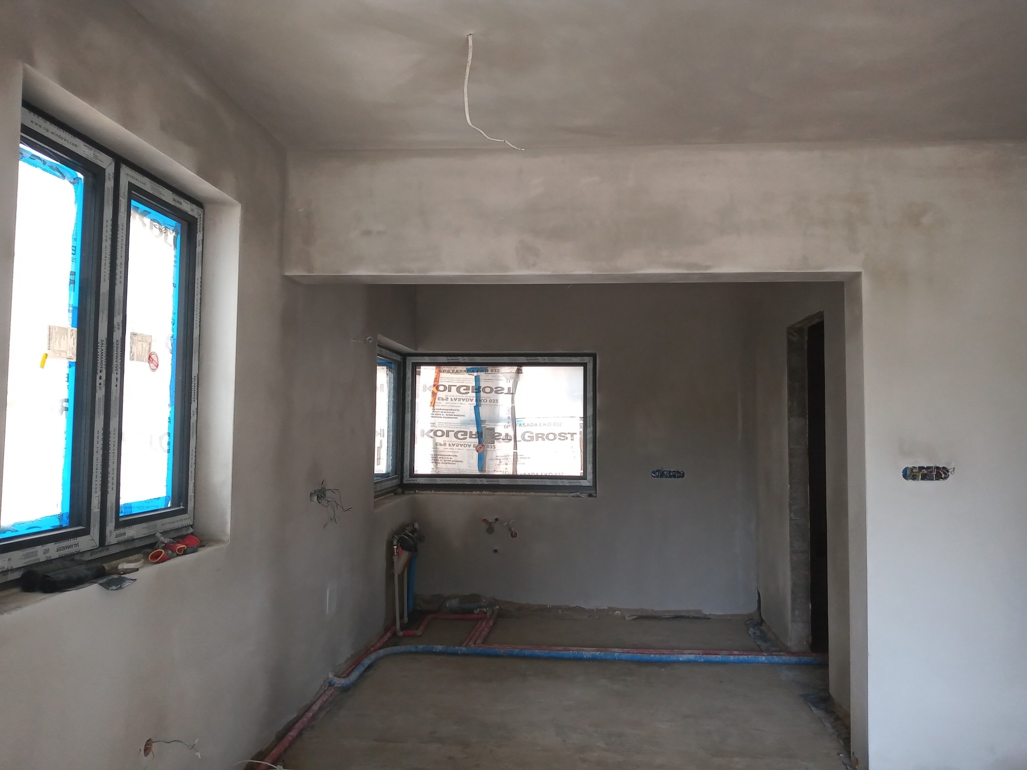 Surowe wnętrze po tynkowaniu maszynowym, widoczne okna z folią ochronną, instalacje wodne i elektryczne w ścianach, sufit z wystającym przewodem.