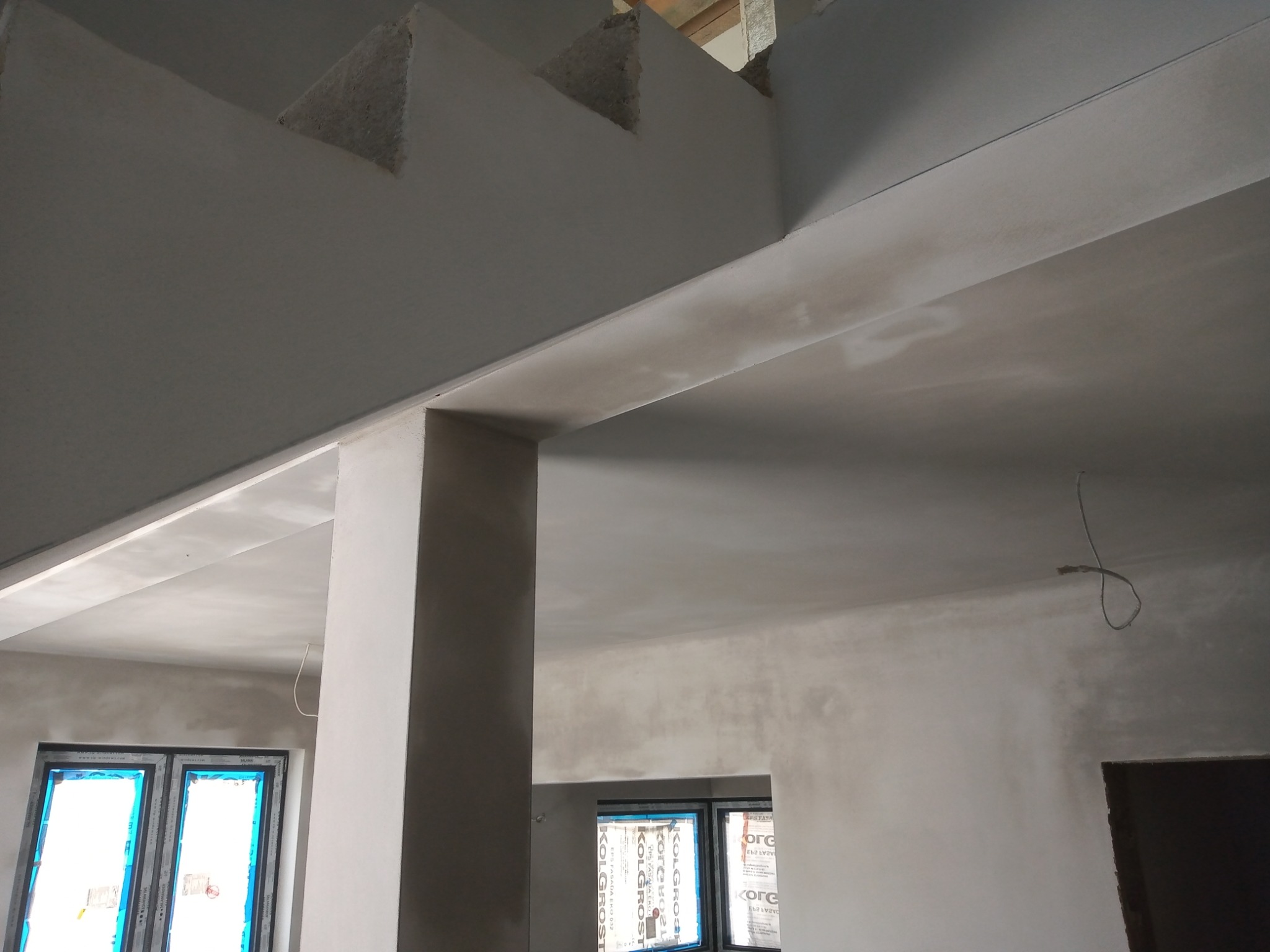 Surowe, szare wnętrze w trakcie prac wykończeniowych; widoczne świeżo otynkowane ściany, kolumny i fragmenty stropu z niedoskonałościami faktury, okna z folią ochronną i wystające kable.