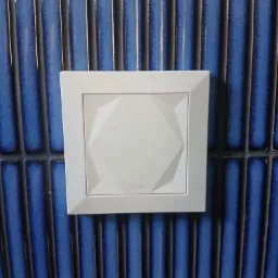 Biały, kwadratowy panel sterowania Loxone zamontowany na ścianie wyłożonej błyszczącymi, pionowymi, niebieskimi płytkami ceramicznymi.