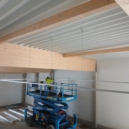 ELEKTRO-INSTAL DARIUSZ DOMARADZKI - Pracownik w żółtej kamizelce na podnośniku koszowym montuje szyny pod sufitem w hali przemysłowej. Widoczne drewniane belki stropowe i metalowe profile podwieszone na linkach.