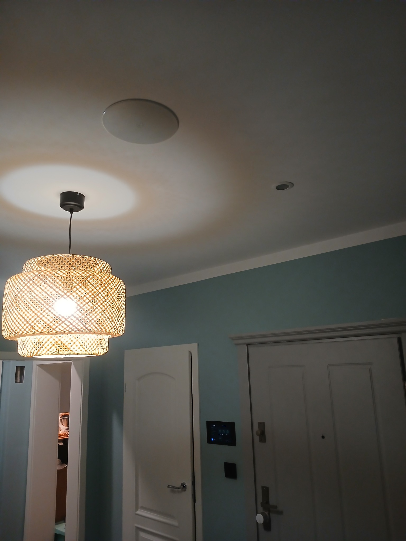 Rattanowy abażur lampy wiszącej, okrągły głośnik sufitowy i panel sterowania Loxone Smart Home na ścianie w tle