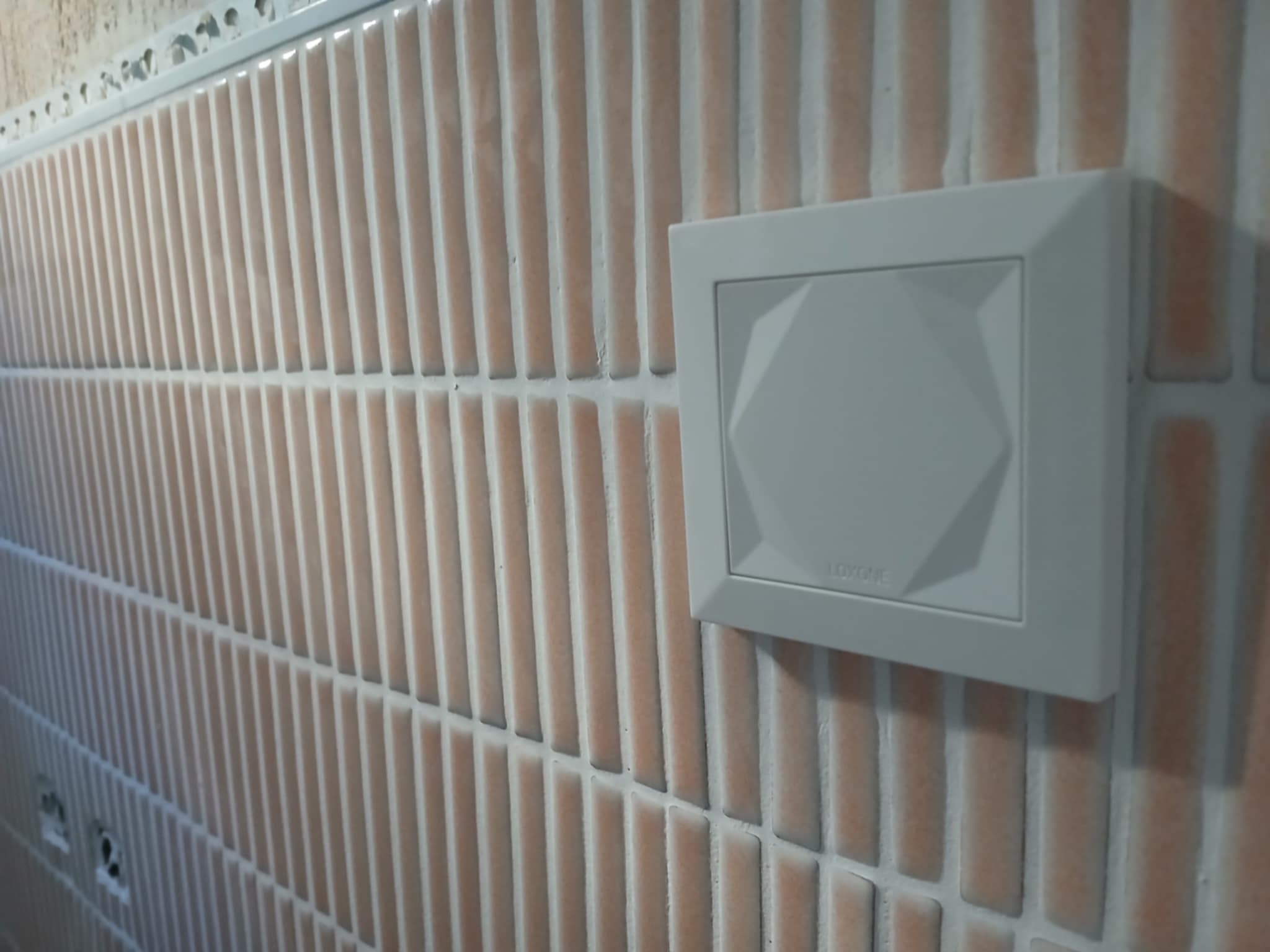 Biały, kwadratowy panel sterujący z geometrycznym wzorem na tle pionowo ułożonych, błyszczących, wąskich, beżowo-brzoskwiniowych płytek ceramicznych w łazience.