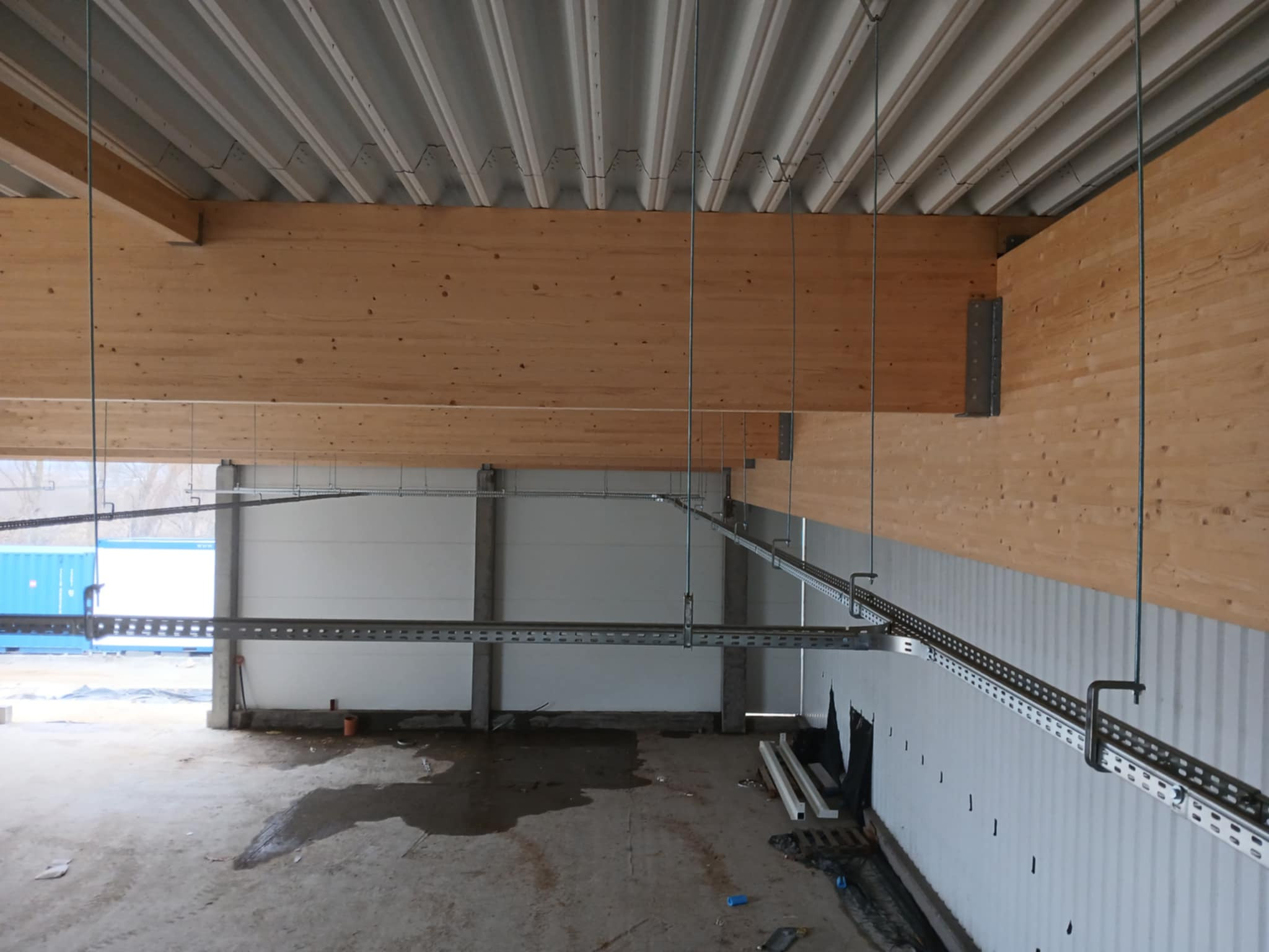 Wnętrze hali produkcyjnej w trakcie prac instalacyjnych, widoczne drewniane belki stropowe, metalowe koryta kablowe podwieszone do sufitu oraz betonowa posadzka w stanie surowym.