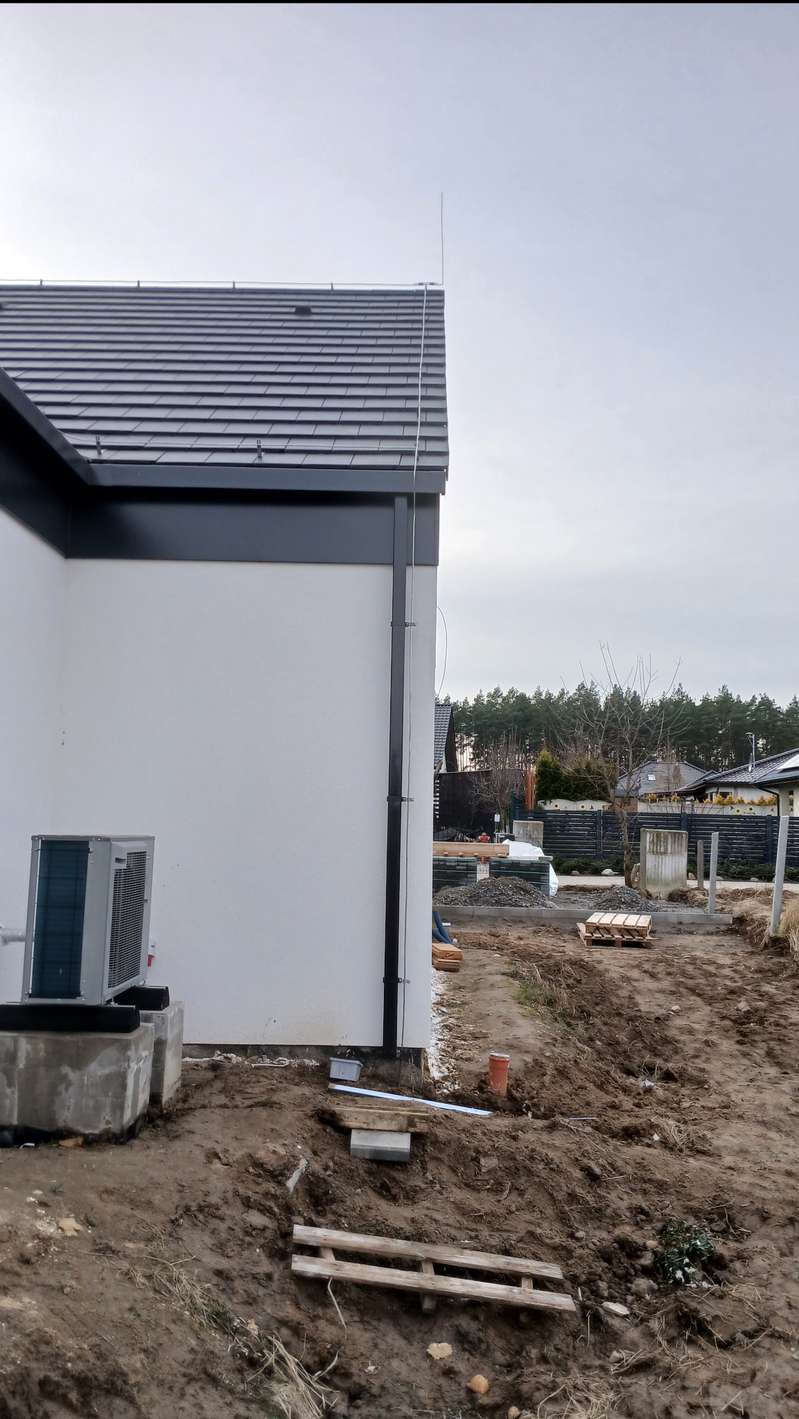 Boczny widok domu z zainstalowaną instalacją odgromową, widoczna metalowa rura na dachu i wzdłuż ściany, oraz jednostka zewnętrzna klimatyzacji na betonowym fundamencie.