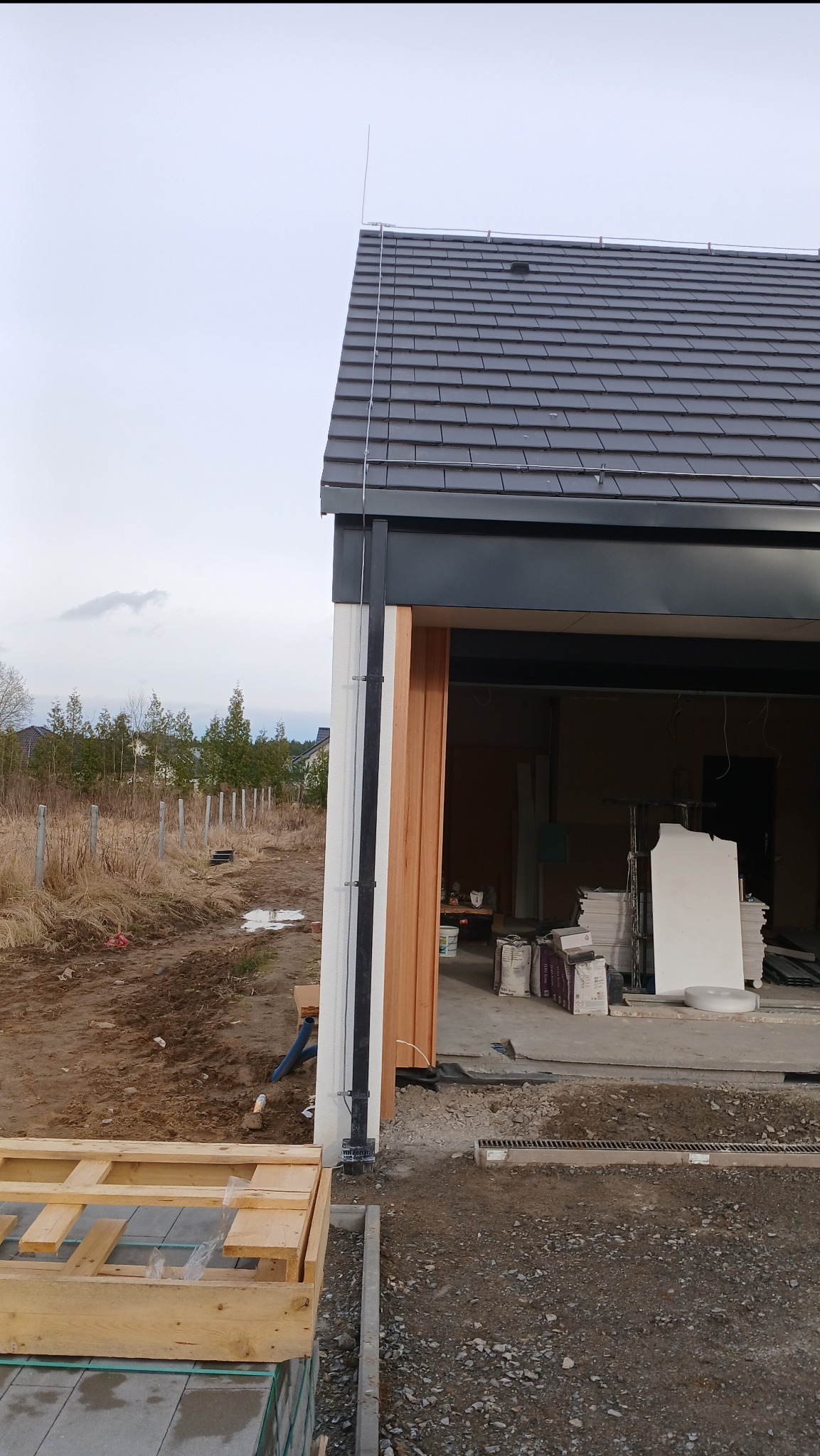 Niedokończony budynek z zamontowaną instalacją odgromową na dachu, widoczne przewody i maszt uziemiający, teren wokół w trakcie prac ziemnych, paleta z kostką brukową w pierwszym planie.