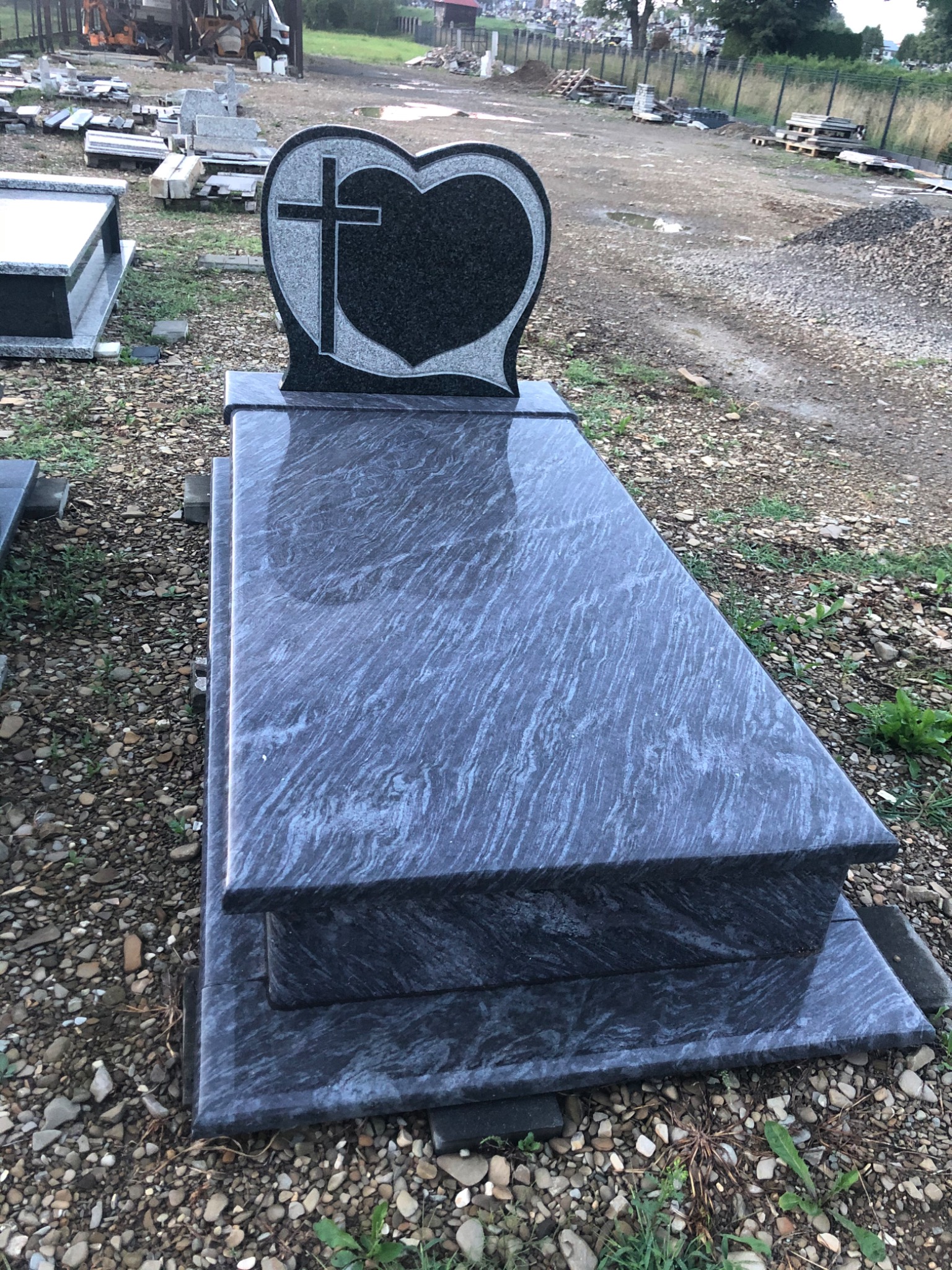 Granitowy pomnik z sercem i krzyżem, widoczne szlifowane krawędzie i polerowana powierzchnia, na tle placu z kamieniami.