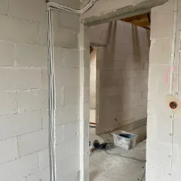 Wnętrze surowego budynku z białych bloczków, widoczne pionowe i poziome rury instalacyjne przymocowane do ścian, w tle narzędzia i przezroczysty pojemnik.