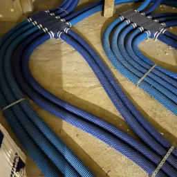 Instalacja rozdzielacza z niebieskich rur karbowanych w szarej, surowej przestrzeni podłogi, widoczne metalowe obejmy i elementy mocujące.