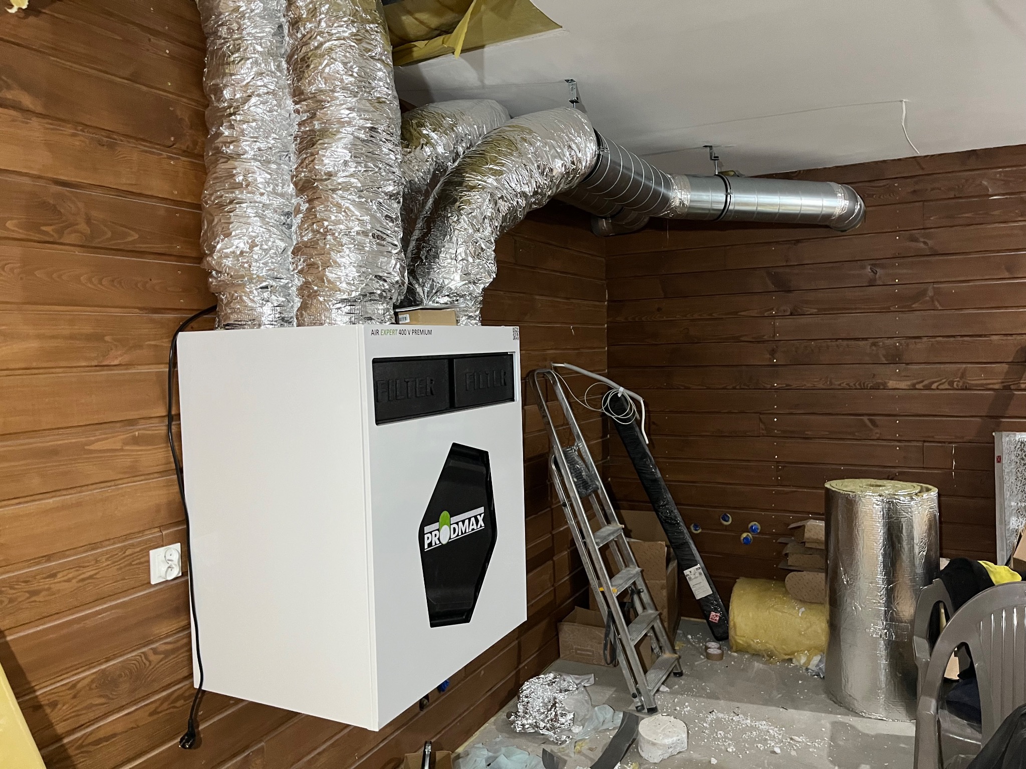 Biała jednostka rekuperacyjna PRODMAX z podłączonymi, srebrnymi przewodami wentylacyjnymi w pomieszczeniu wykończonym drewnianą boazerią. Widoczna drabina i materiały izolacyjne.