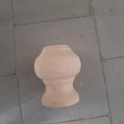 Surowy, niedokończony element toczony z jasnego drewna, przypominający ozdobną nóżkę meblową, leżący na szarych płytkach chodnikowych.