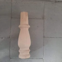 Surowa, drewniana noga meblowa o toczonym kształcie, leżąca na szarej, betonowej posadzce.