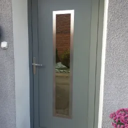 Szare drzwi zewnętrzne z podłużnym, metalowym przeszkleniem, zamontowane w elewacji z szarą strukturą, widoczne różowe kwiaty po prawej stronie, odbicie ceglanego budynku w szybie.