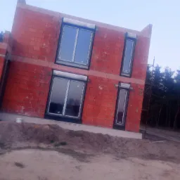 Surowy budynek w trakcie budowy z zamontowanymi oknami z czarnymi ramami i częściowo opuszczonymi roletami zewnętrznymi, widoczne bloczki ceramiczne i ziemia.