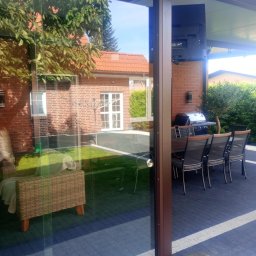 Rafał Graczyk Rafmont - Przesuwne szklane drzwi tarasowe z brązowymi ramami, otwierające się na patio z grillem i meblami ogrodowymi. Widoczny budynek w tle.