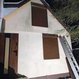 Rafał Graczyk Rafmont - Dom z brązowymi roletami zewnętrznymi na oknach, widziany nocą. Aluminiowa drabina oparta o ścianę. Śnieg leży u podstawy budynku.