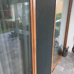 Fragment okna z drewnopodobną ramą i zamontowaną moskitierą, widok na zewnątrz z odbiciem wnętrza, w tle narzędzia i doniczka z kwiatami.