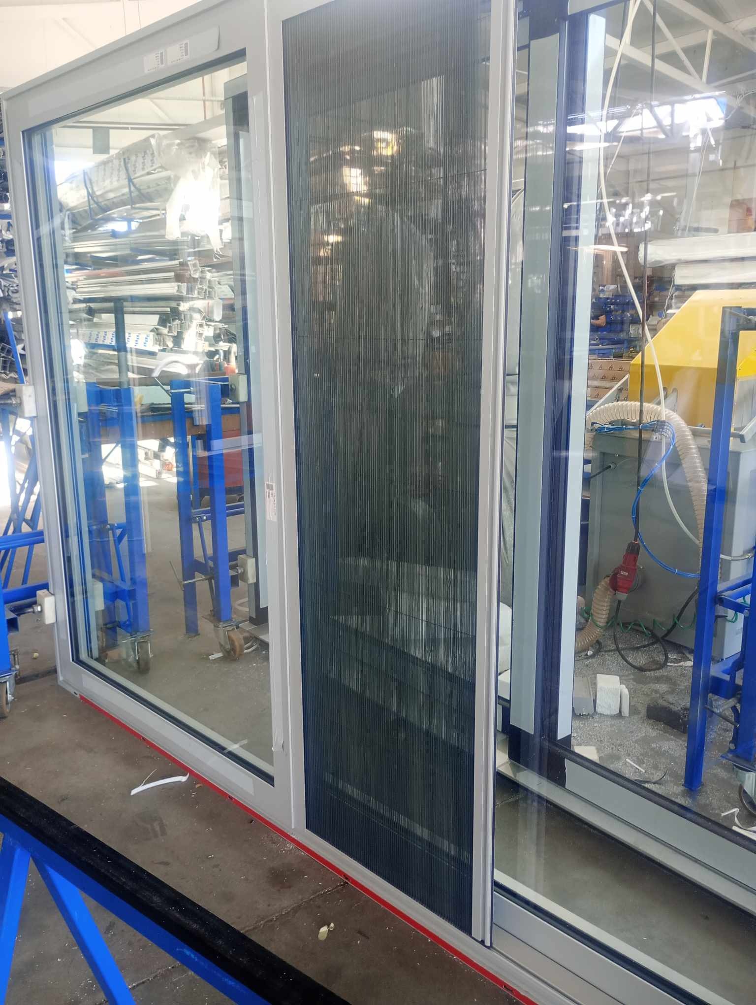 Aluminiowa rama okienna ze zintegrowaną moskitierą plisowaną, widoczna w hali produkcyjnej z maszynami i elementami konstrukcyjnymi w tle.