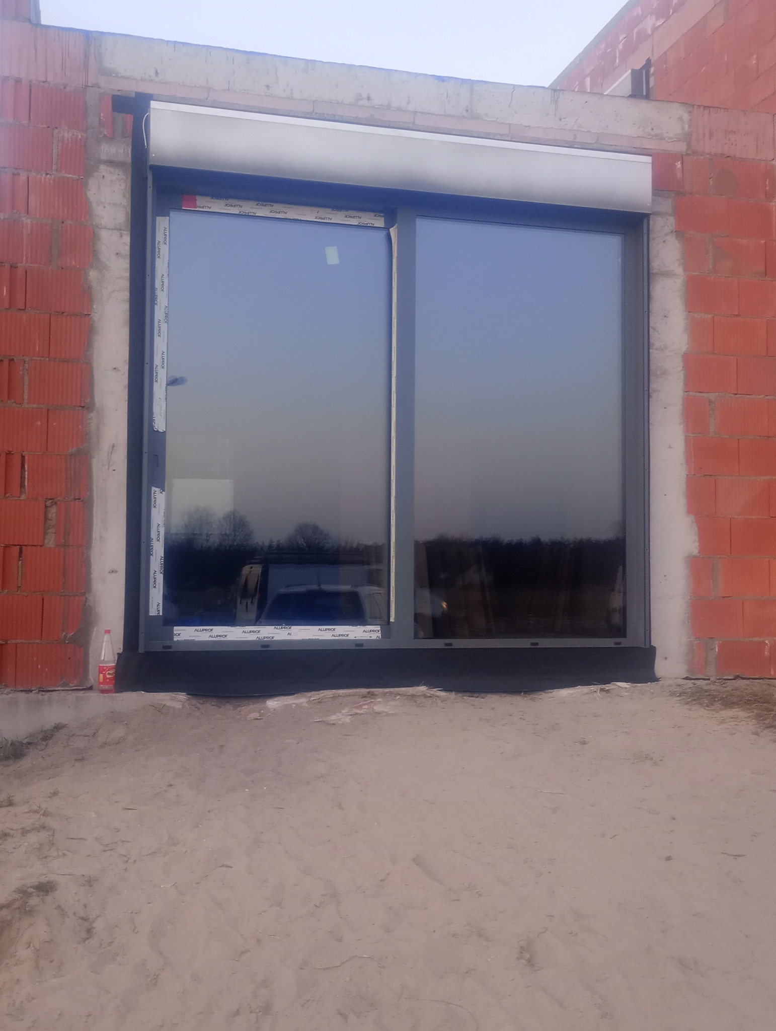 Szare okno PCV z roletą zewnętrzną zamontowane w surowym murze z czerwonej cegły, widoczne naklejki producenta na ramie okiennej i butelka po napoju obok.