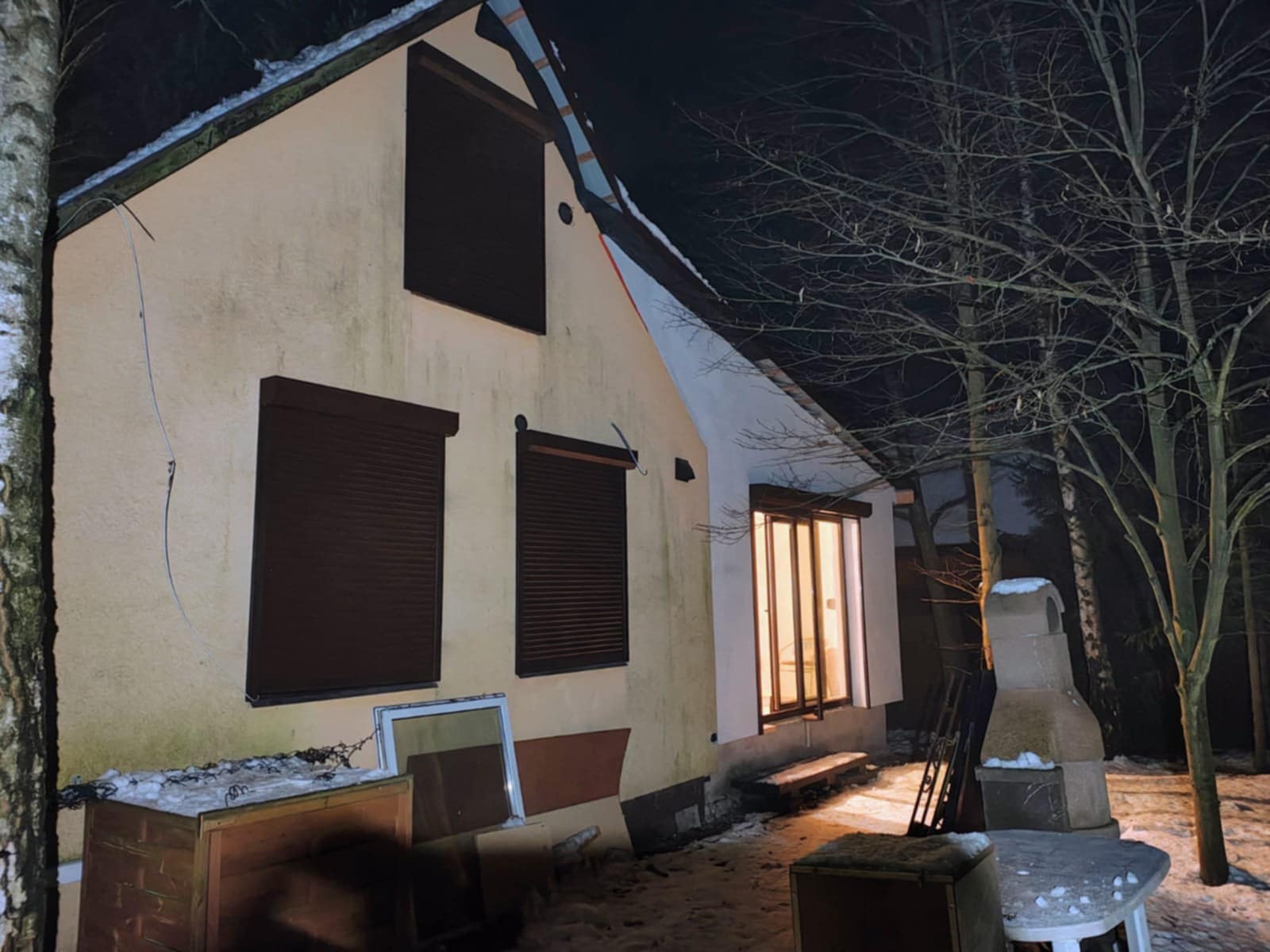 Dom jednorodzinny z brązowymi roletami zewnętrznymi na oknach, sfotografowany nocą. Widoczne drzewa i elementy otoczenia domu.