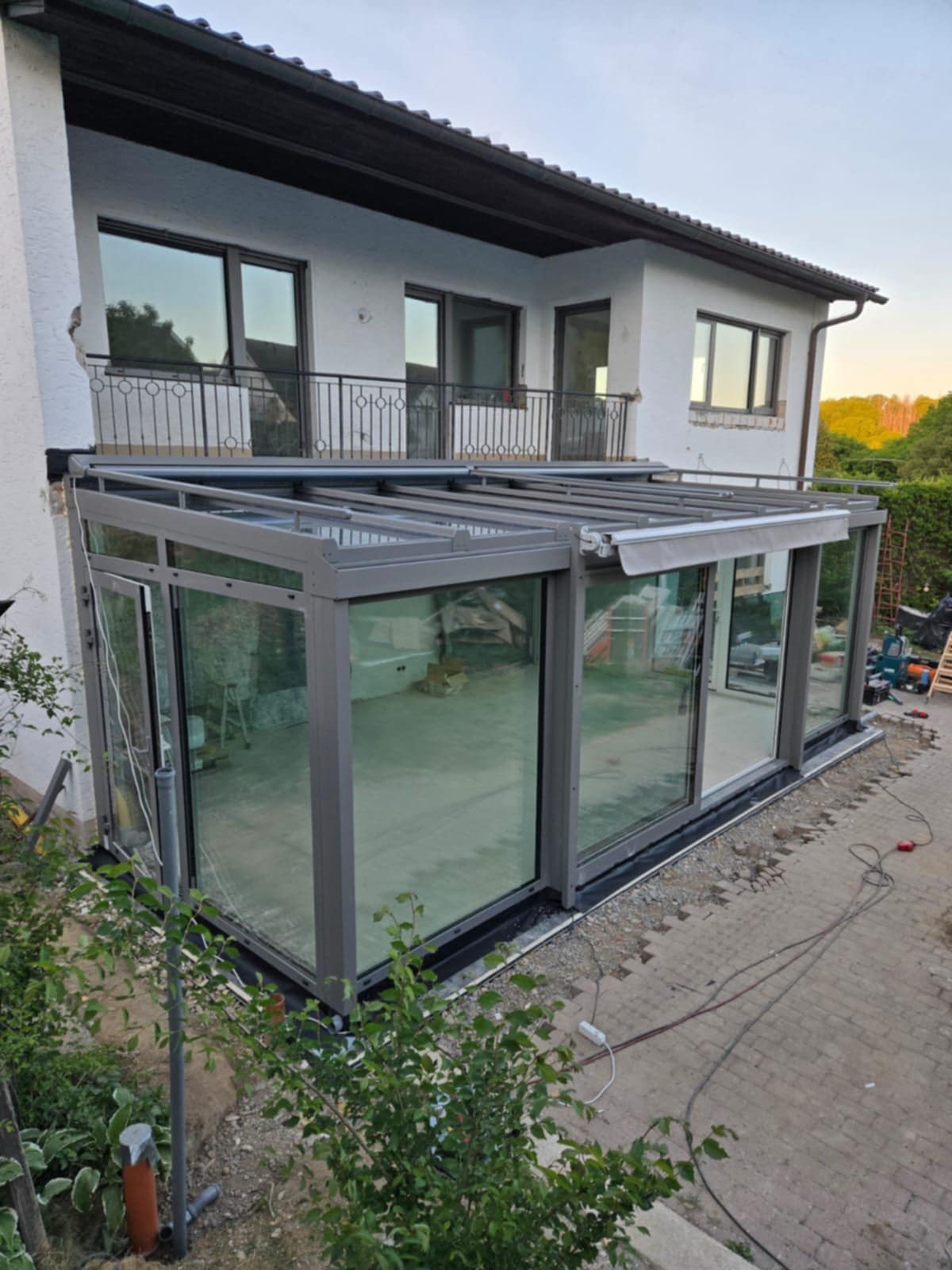 Szklana zabudowa tarasu z szarymi ramami, widok z zewnątrz, częściowo zwinięta markiza, biały budynek w tle, kostka brukowa na pierwszym planie.