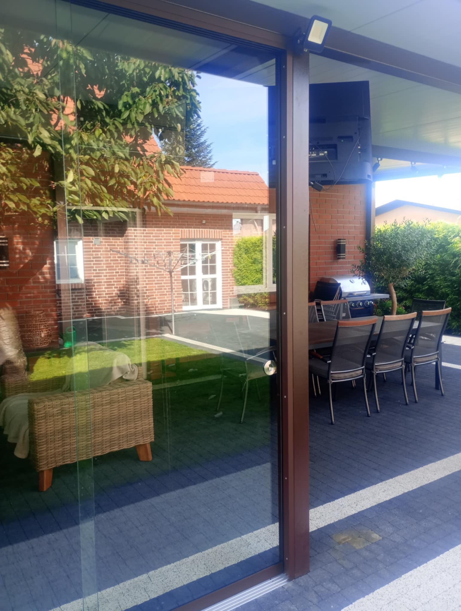 Przesuwne szklane drzwi tarasowe z brązowymi ramami, otwierające się na patio z grillem i meblami ogrodowymi. Widoczny budynek w tle.