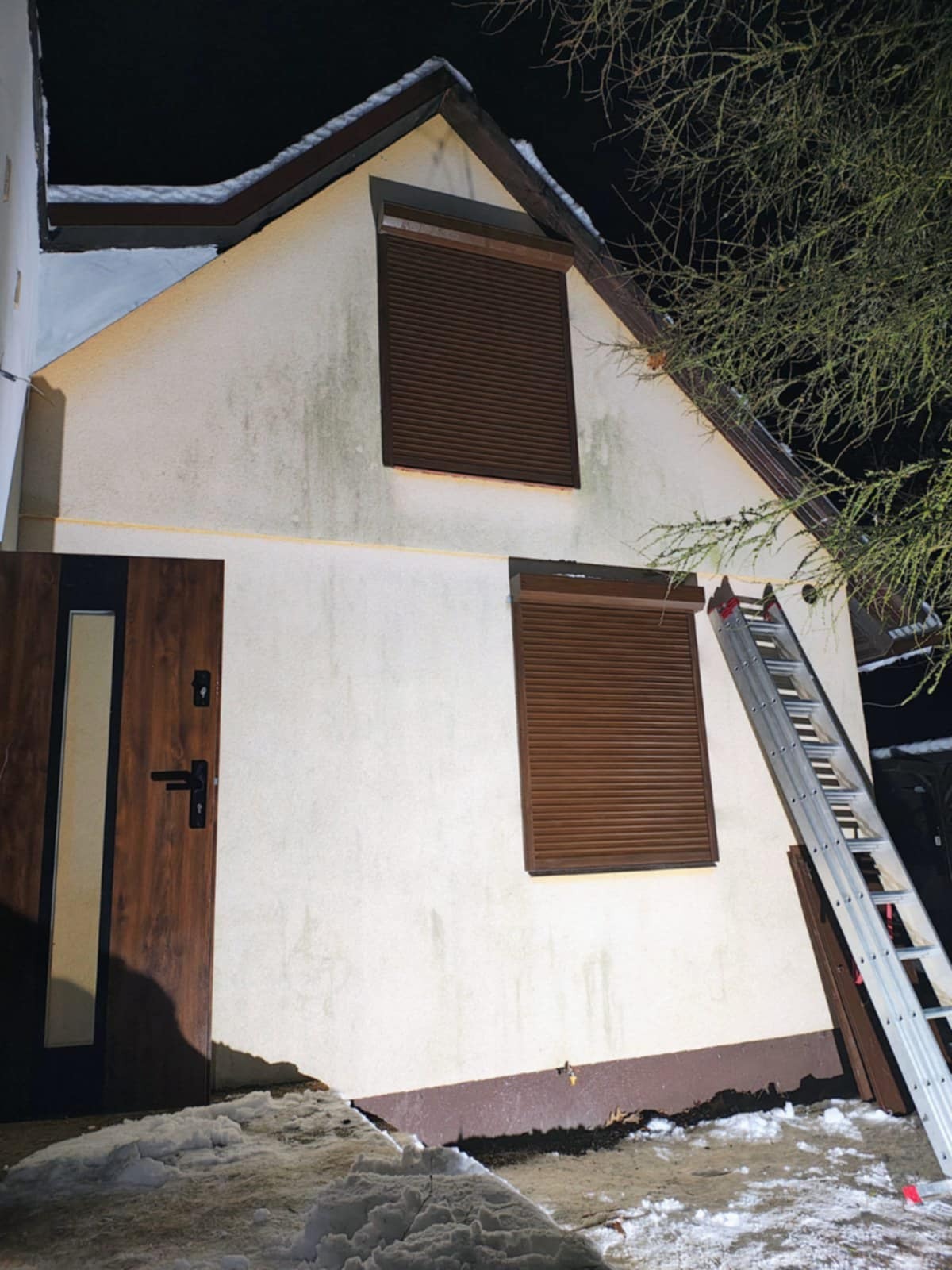 Dom z brązowymi roletami zewnętrznymi na oknach, widziany nocą. Aluminiowa drabina oparta o ścianę. Śnieg leży u podstawy budynku.