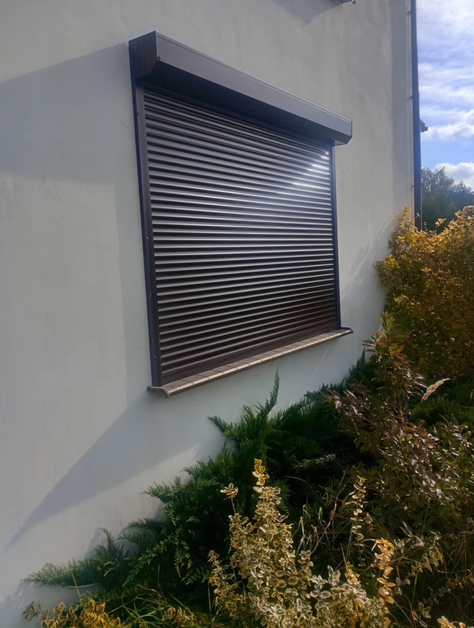 Brązowa roleta zewnętrzna zamontowana na oknie budynku z jasną elewacją, otoczona zielenią. Widoczne detale montażu i kolorystyka dopasowana do otoczenia.