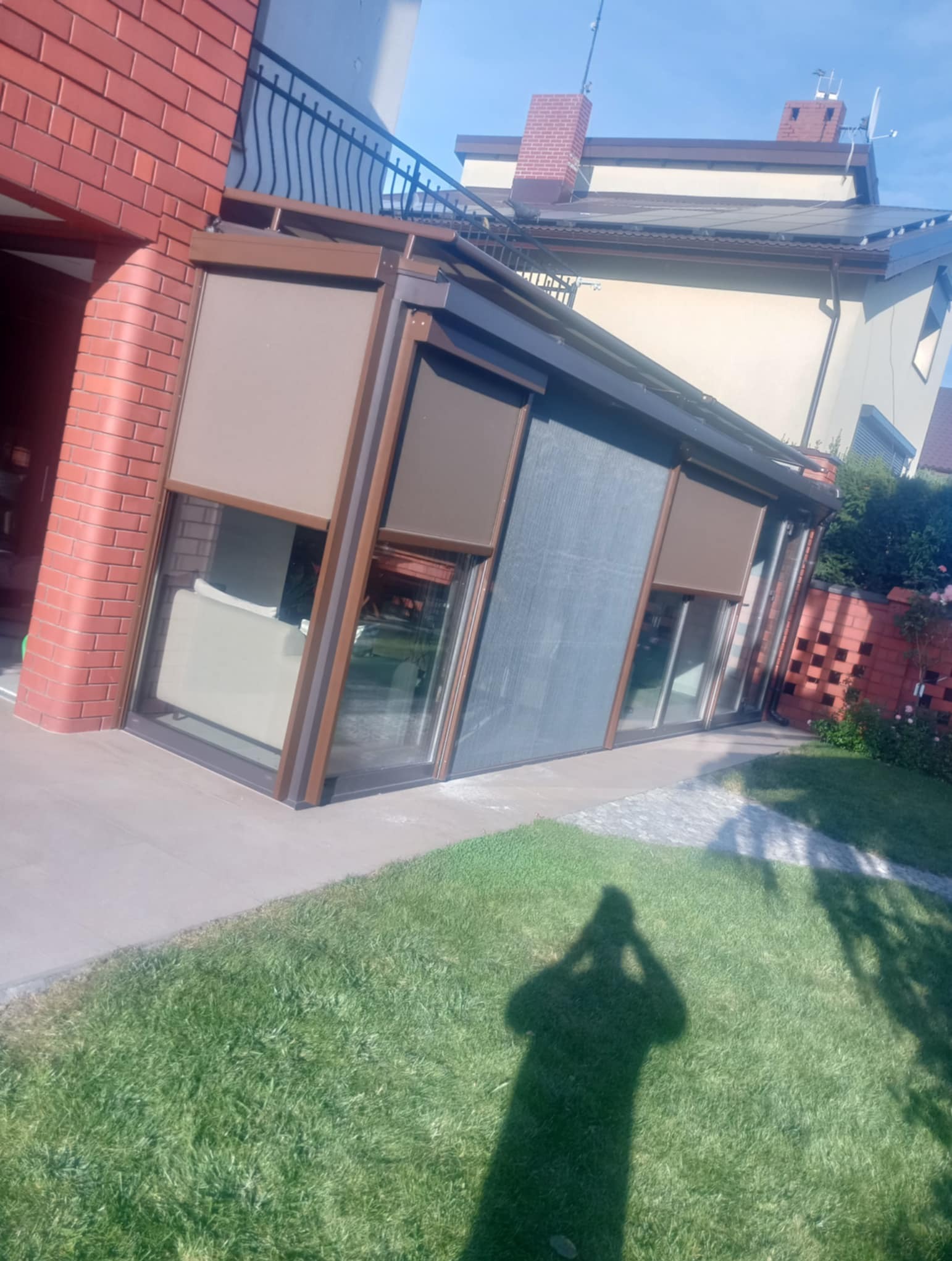 Zadaszenie tarasu z brązowymi ramami i częściowo opuszczonymi roletami w odcieniach beżu i szarości, widoczne z zewnątrz na tle domu i trawnika.