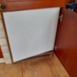 AgaSprząta - Otwarta szafka kuchenna z pustą przestrzenią wewnątrz, widoczne zawiasy i fragmenty płytek ceramicznych na ścianie i podłodze.