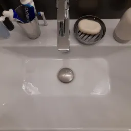 Biała umywalka z chromowaną baterią, mydłem w kostce, dozownikiem i szczoteczkami do zębów. Widoczne krople wody, sugerujące świeże umycie.