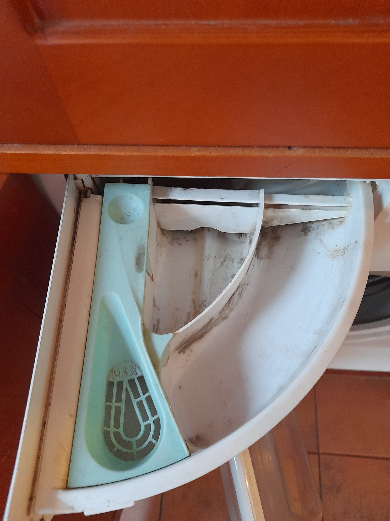 Z bliska: szuflada na detergent w pralce, z widocznymi zabrudzeniami i osadami. Elementy plastikowe w kolorze bieli i seledynu, wnętrze szuflady pokryte ciemnym nalotem.