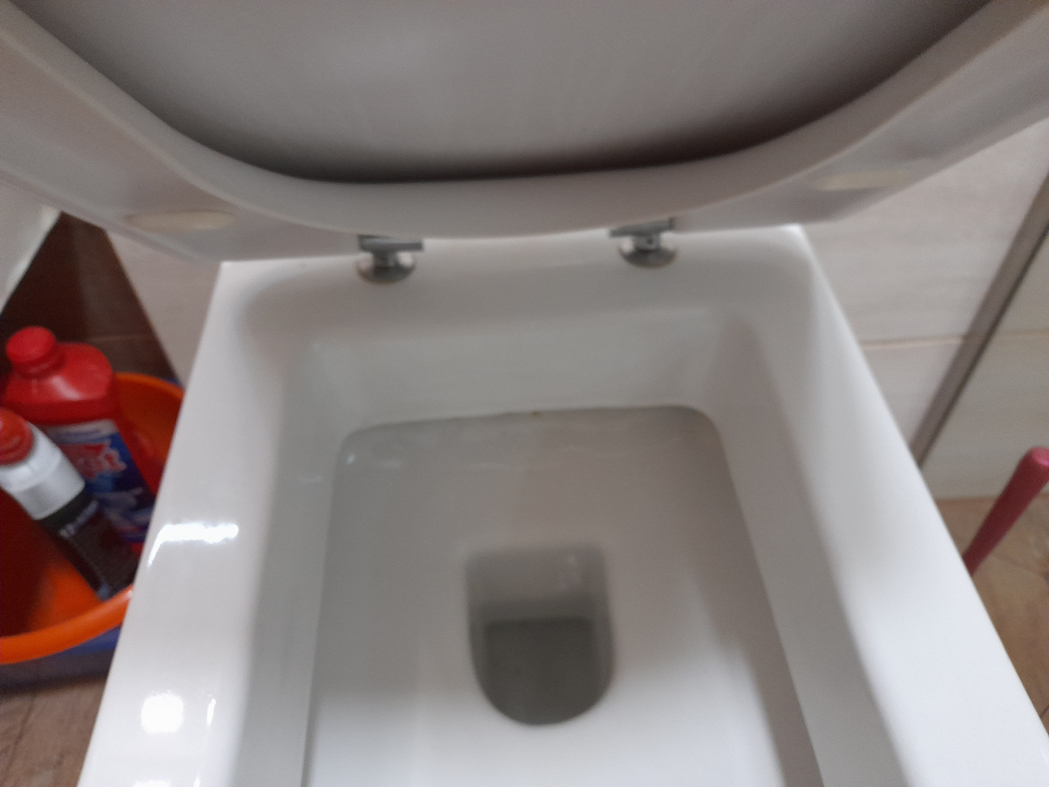 Biała muszla klozetowa z podniesioną deską, widok z góry. W tle wiadro z detergentami i szczotka. Ukazanie czystości po sprzątaniu toalety.