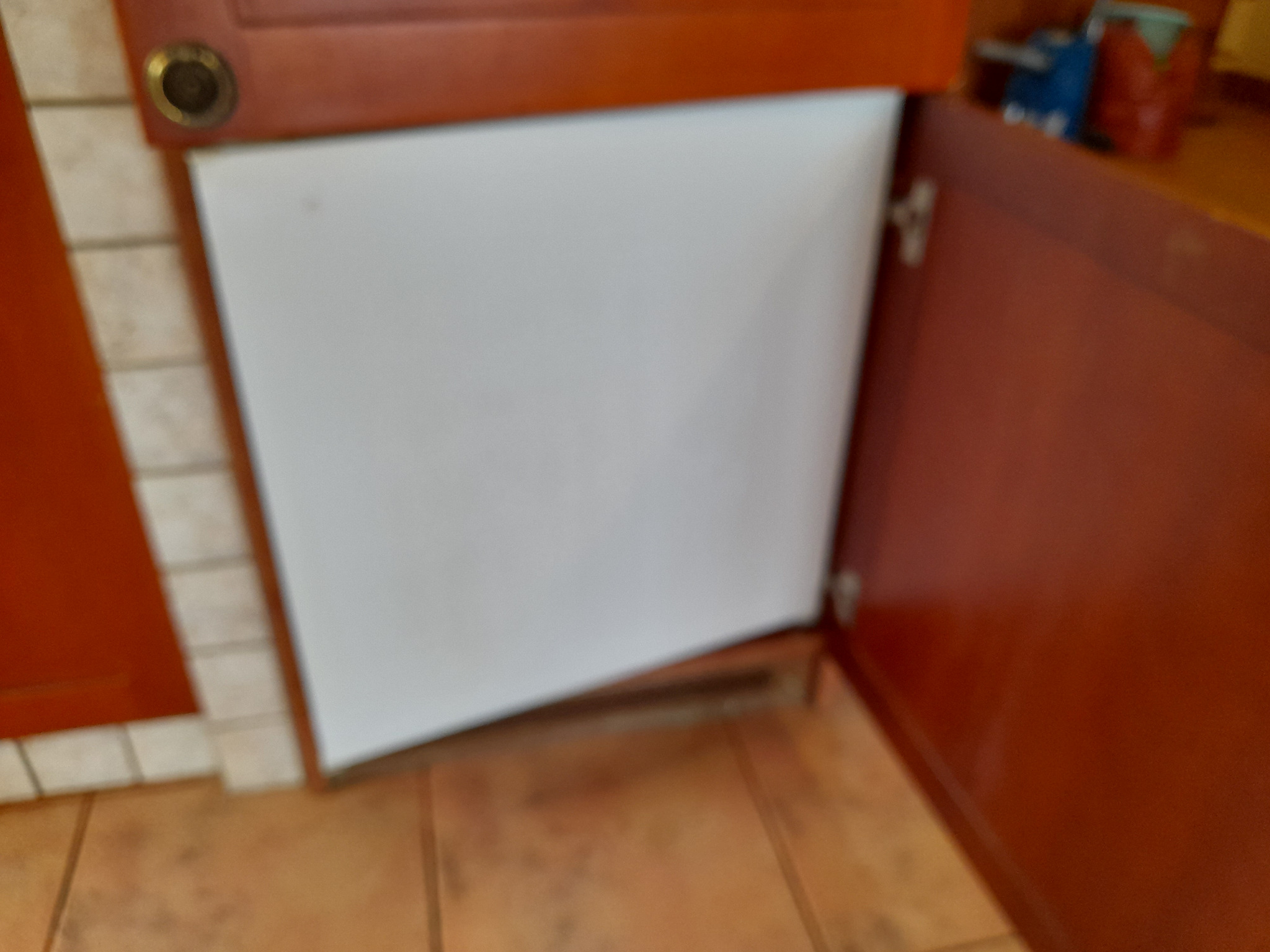 Otwarta szafka kuchenna z pustą przestrzenią wewnątrz, widoczne zawiasy i fragmenty płytek ceramicznych na ścianie i podłodze.