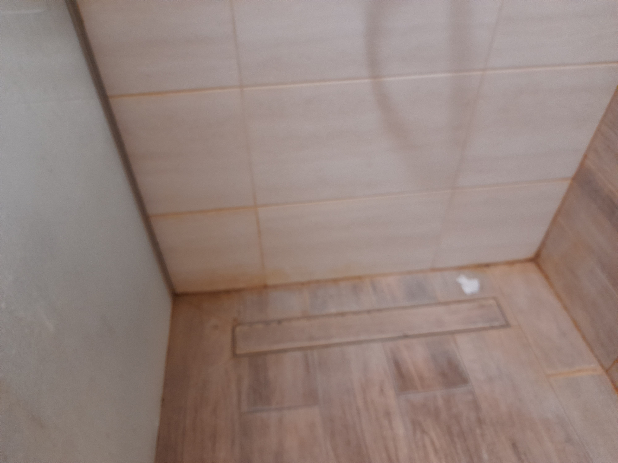 Narożnik łazienki z brązowymi płytkami imitującymi drewno na podłodze i ścianie, widoczny odpływ liniowy i resztki po sprzątaniu.