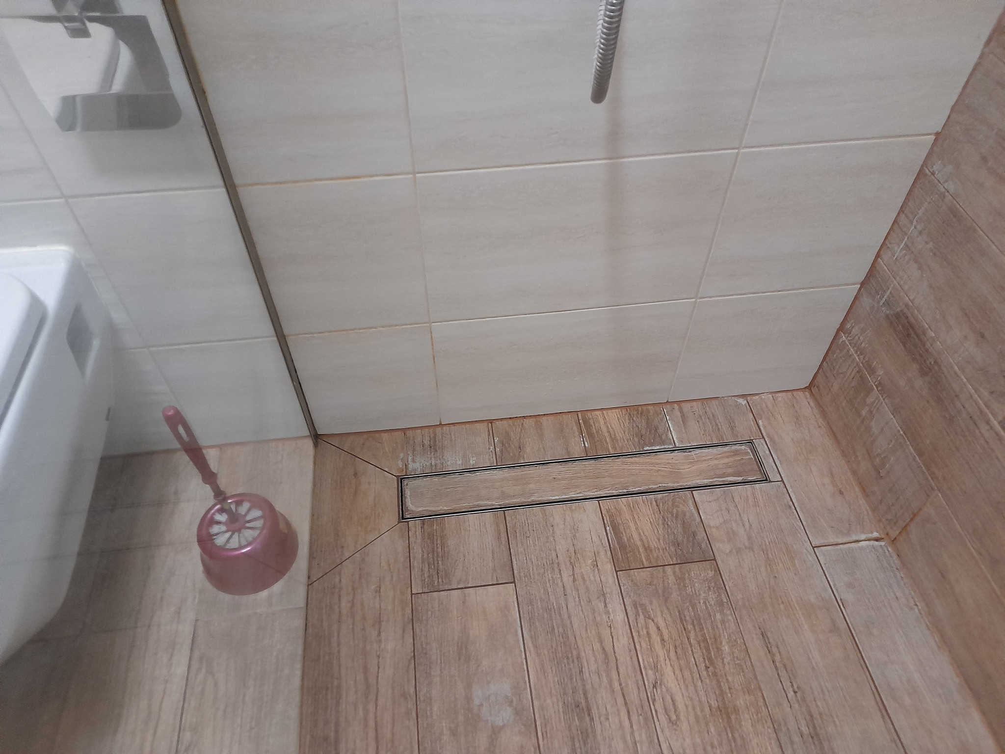 Ukończona łazienka z brązowymi płytkami imitującymi drewno, odpływem liniowym i toaletą. Widoczna szczotka do WC.