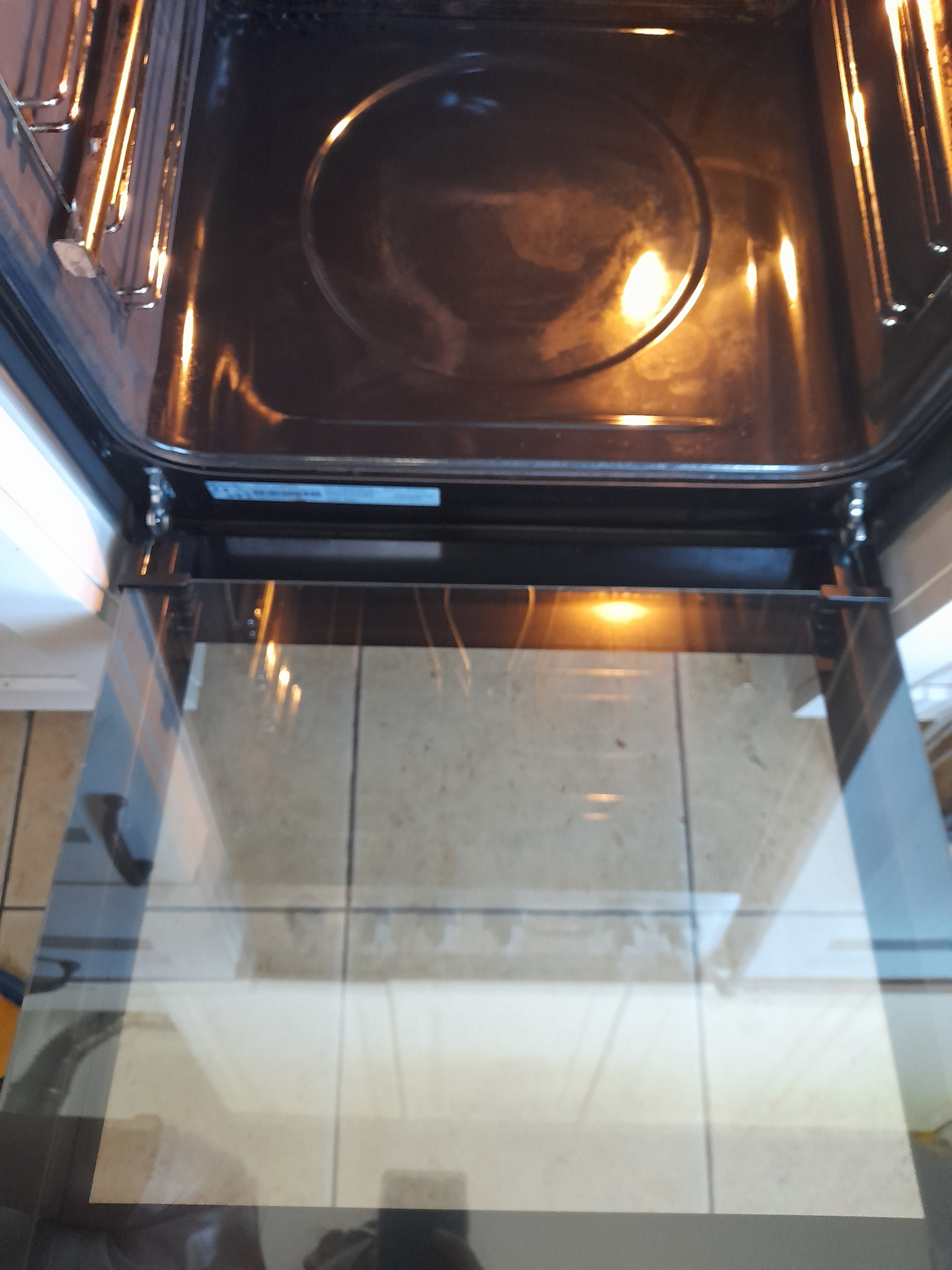 Wnętrze otwartego piekarnika z widocznymi elementami grzejnymi i czystą szybą, odbijającą światło. Widoczne płytki podłogowe w tle.