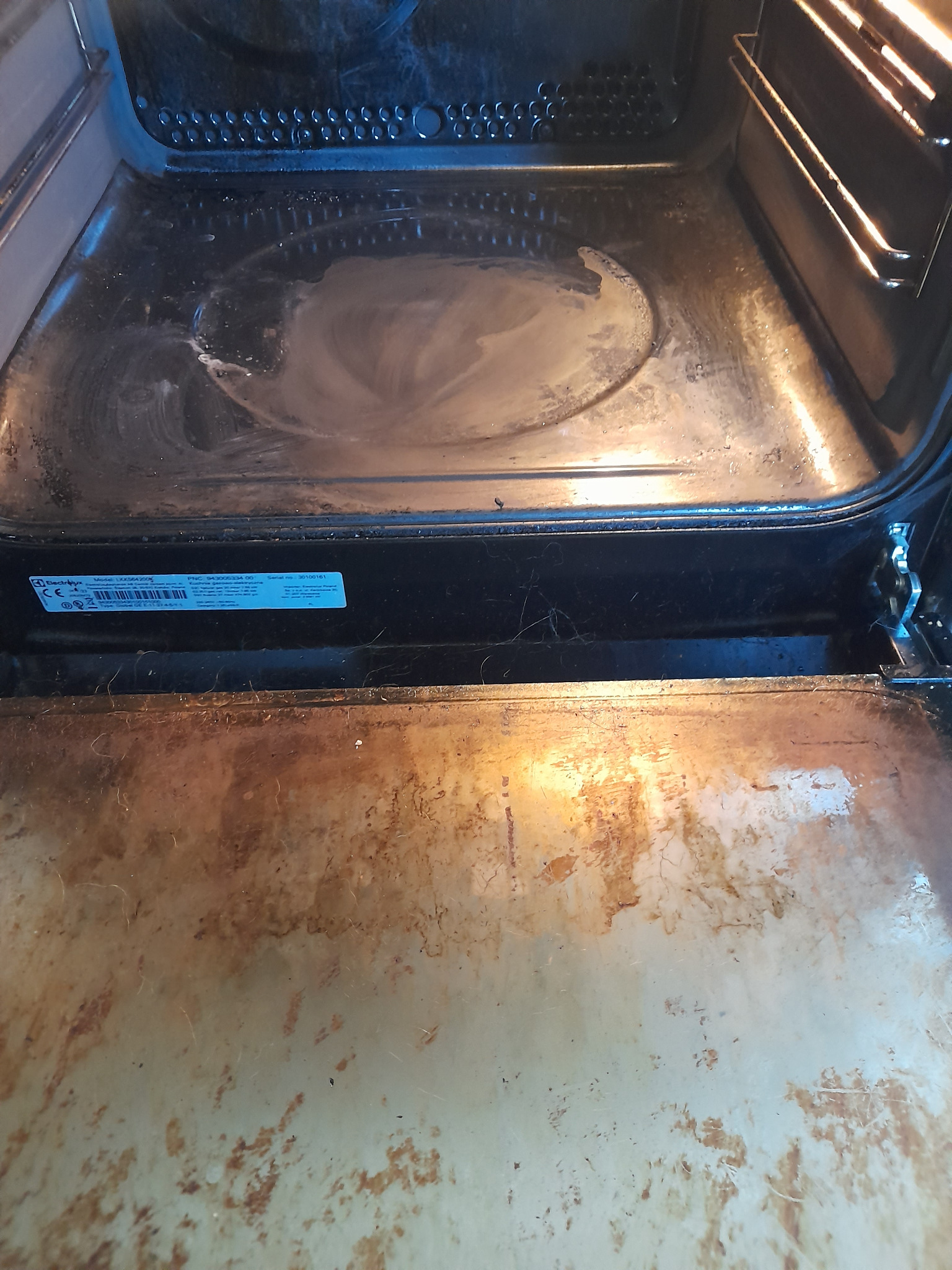 Wnętrze brudnego piekarnika z wysuniętą, mocno zabrudzoną blachą. Widoczne przypalone resztki jedzenia. Potrzebne gruntowne czyszczenie.