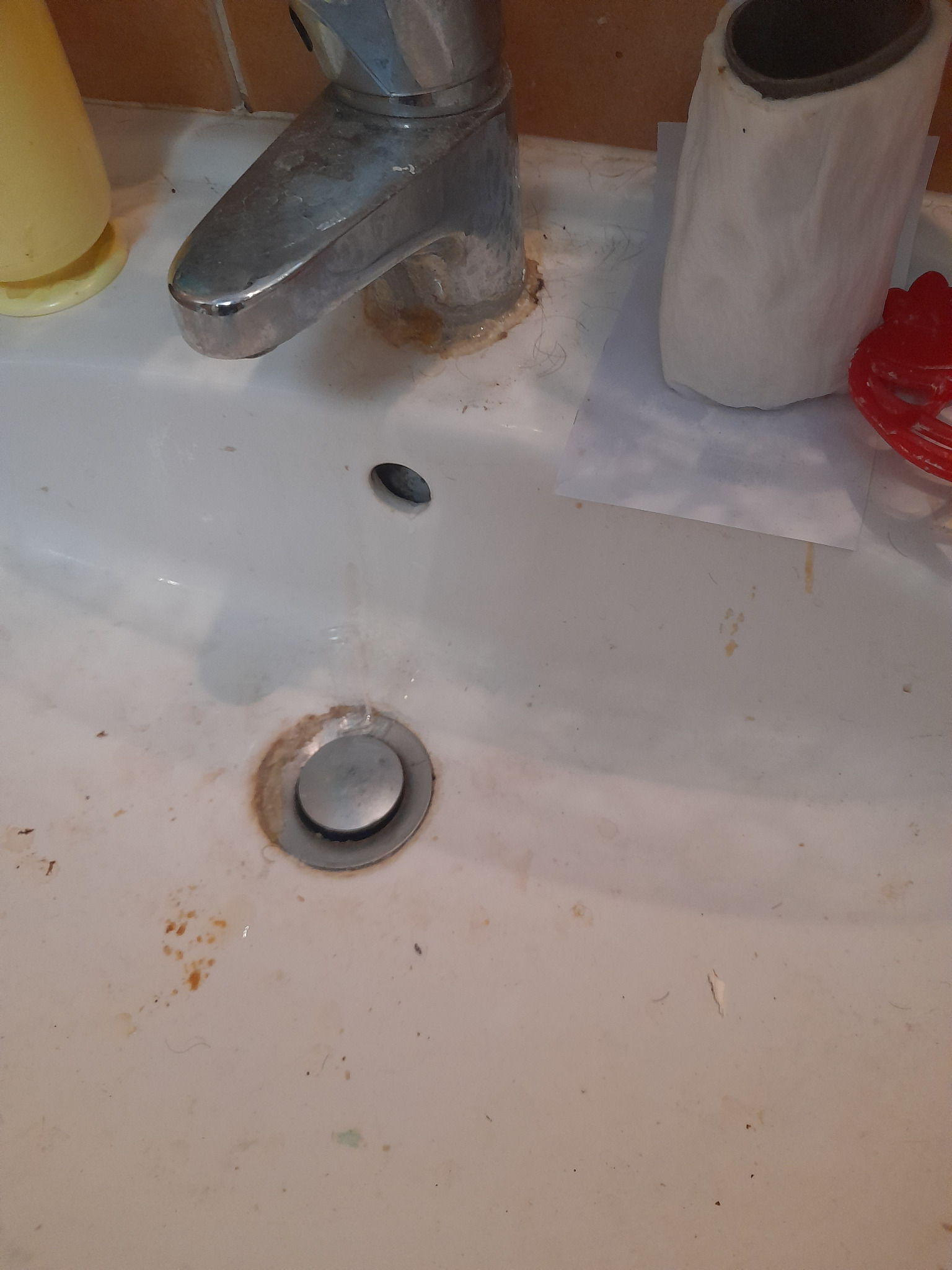 Zanieczyszczona umywalka z osadem i resztkami. Widoczny kran, odpływ i akcesoria łazienkowe. Brak świeżości i czystości.