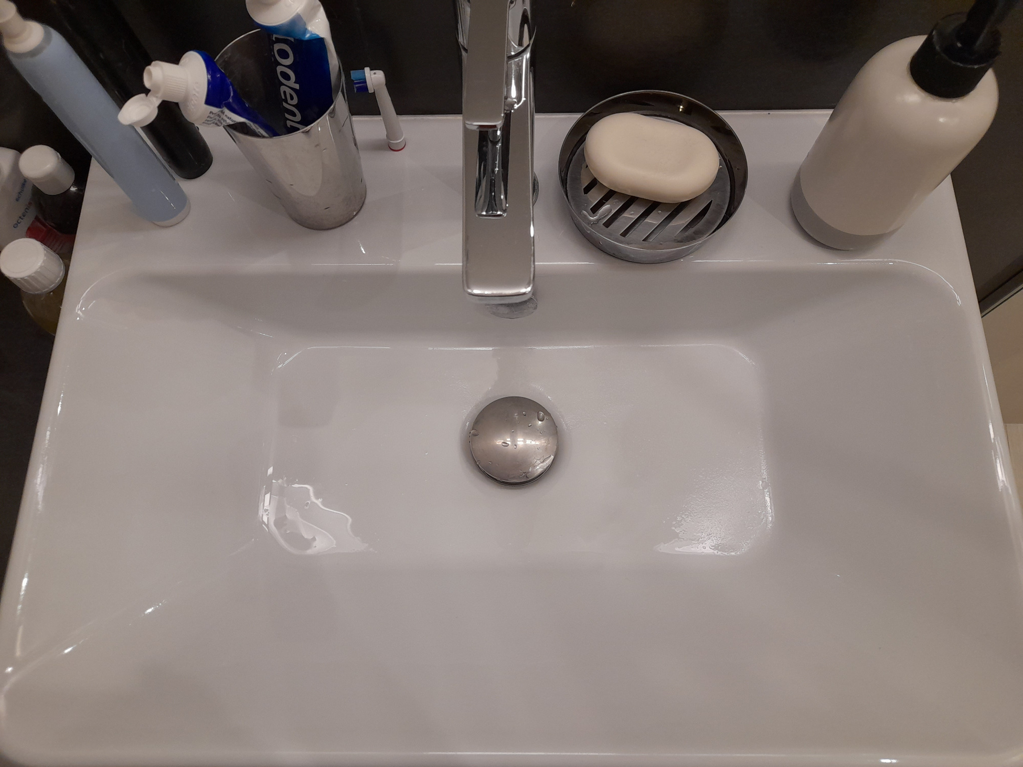 Biała umywalka z chromowaną baterią, mydłem w kostce, dozownikiem i szczoteczkami do zębów. Widoczne krople wody, sugerujące świeże umycie.