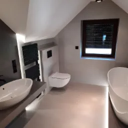 Nowoczesna łazienka na poddaszu z umywalką nablatową, podwieszaną toaletą i wolnostojącą wanną, oświetlona listwami LED wzdłuż podłogi i pod umywalką, oraz czarnym grzejnikiem drabinkowym.