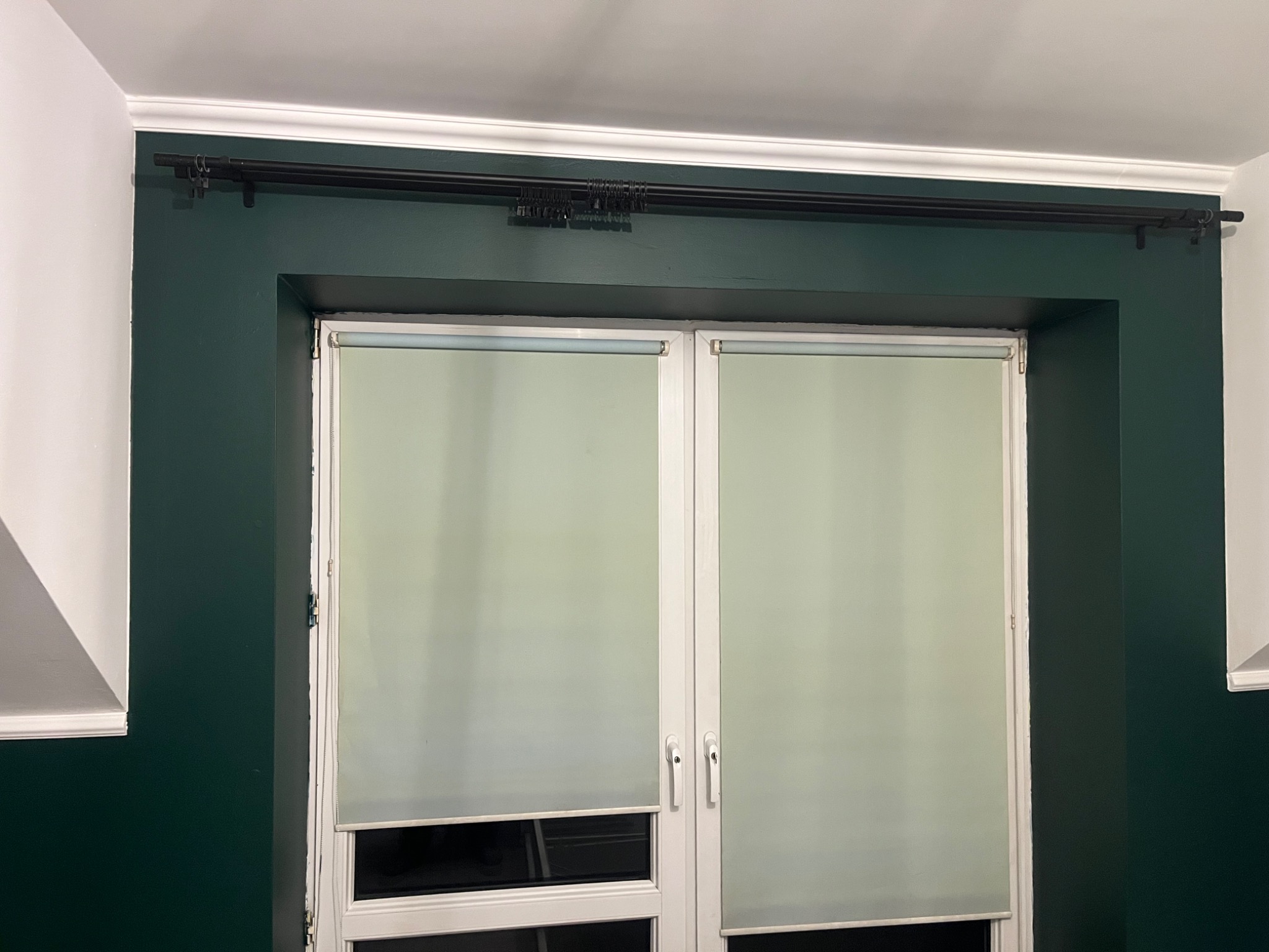 Ściana pomalowana na ciemnozielony kolor z białymi oknami i roletami, karnisz w kolorze czarnym.