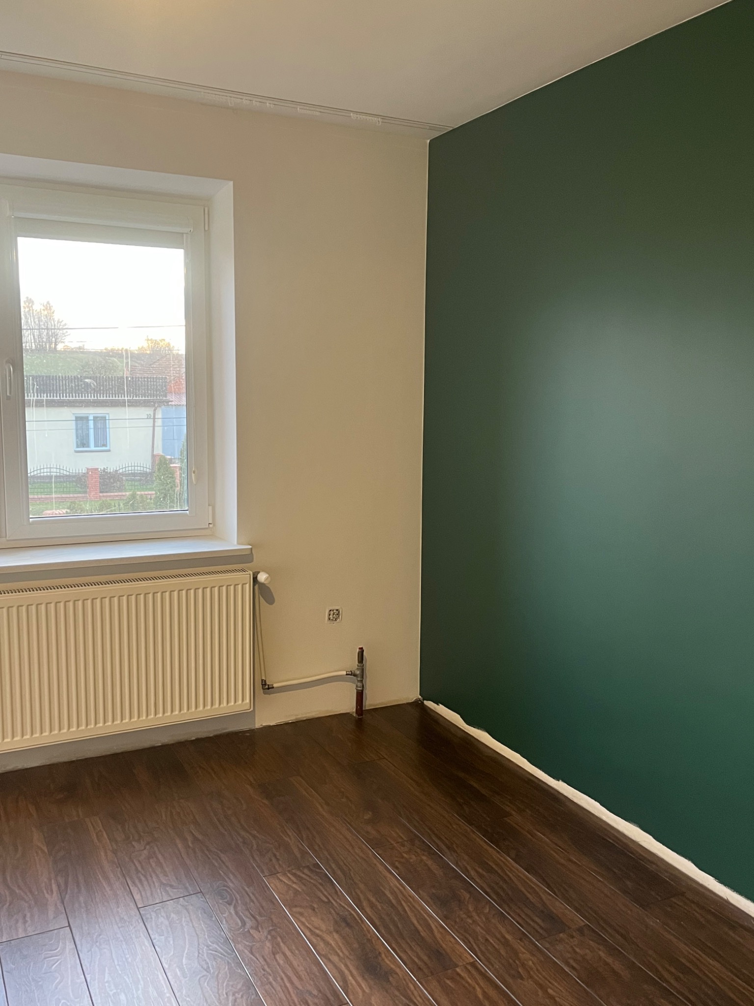 Pomalowany na ciemnozielono fragment ściany w pokoju z panelami podłogowymi, widoczne okno z grzejnikiem i białą ścianą obok.
