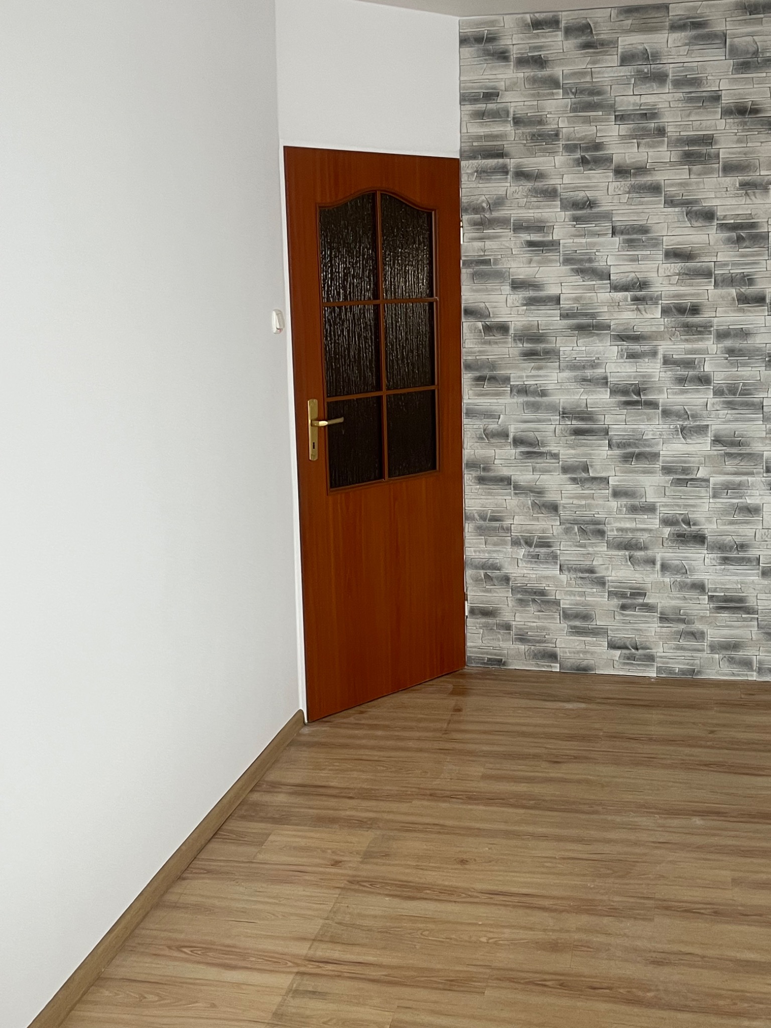 Wykończone wnętrze: drewnopodobna podłoga, biała ściana i ściana pokryta szarą okładziną imitującą kamień, widoczne drewniane drzwi z szybą.